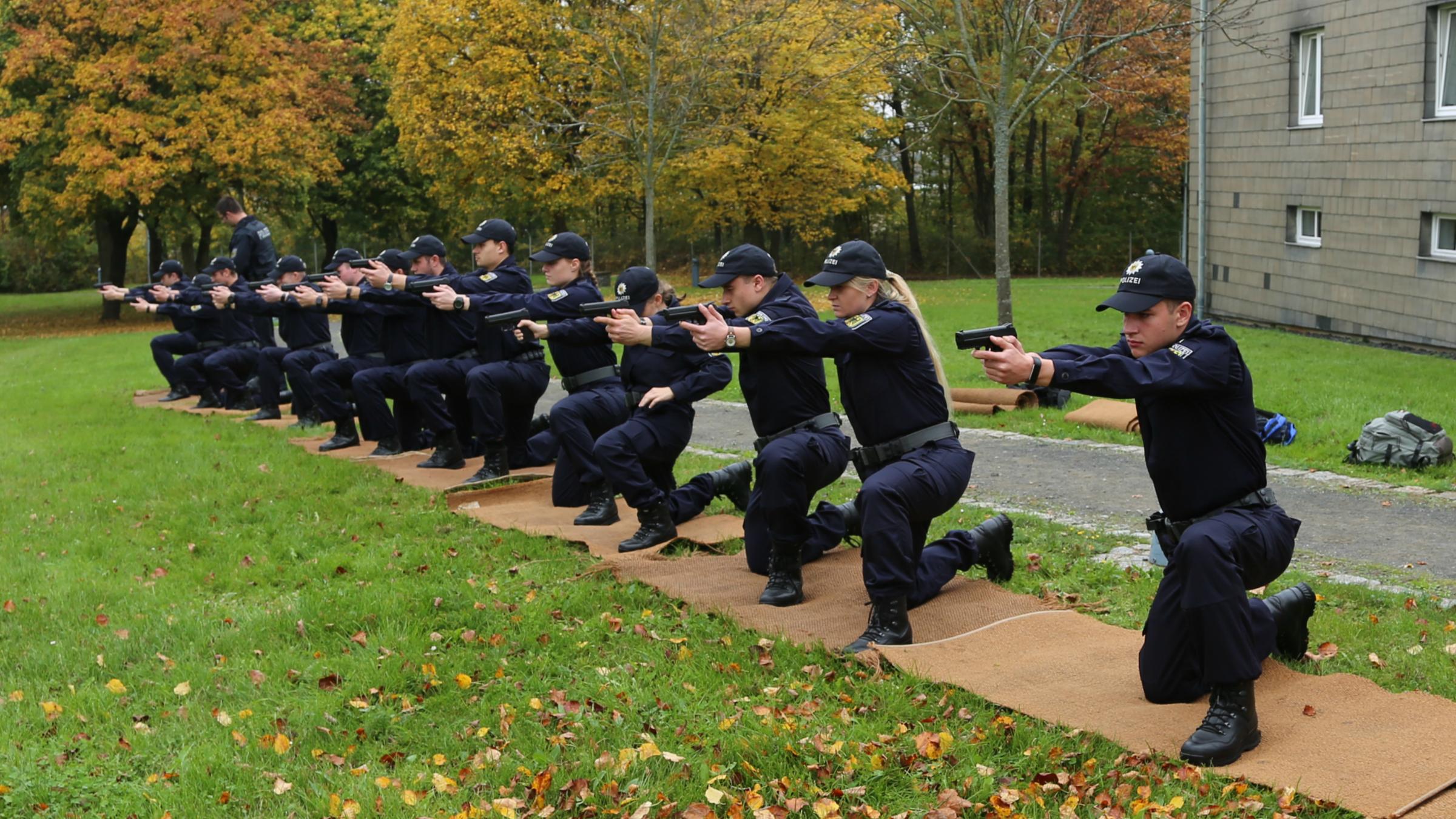 "ZDF.reportage - Nachwuchs für die Bundespolizei - Zwischen Schulbank und Schießstand": Eine Gruppe von 13 Personen in Einsatzanzügen der Bundespolizei kniet nebeneinander auf braunen Matten auf einer Wiese und zielt mit ihren Waffen geradeaus. Ein Mann in gleicher Kleidung steht hinter der Person am äußersten Bildrand und begutachtet die Waffenhaltung.