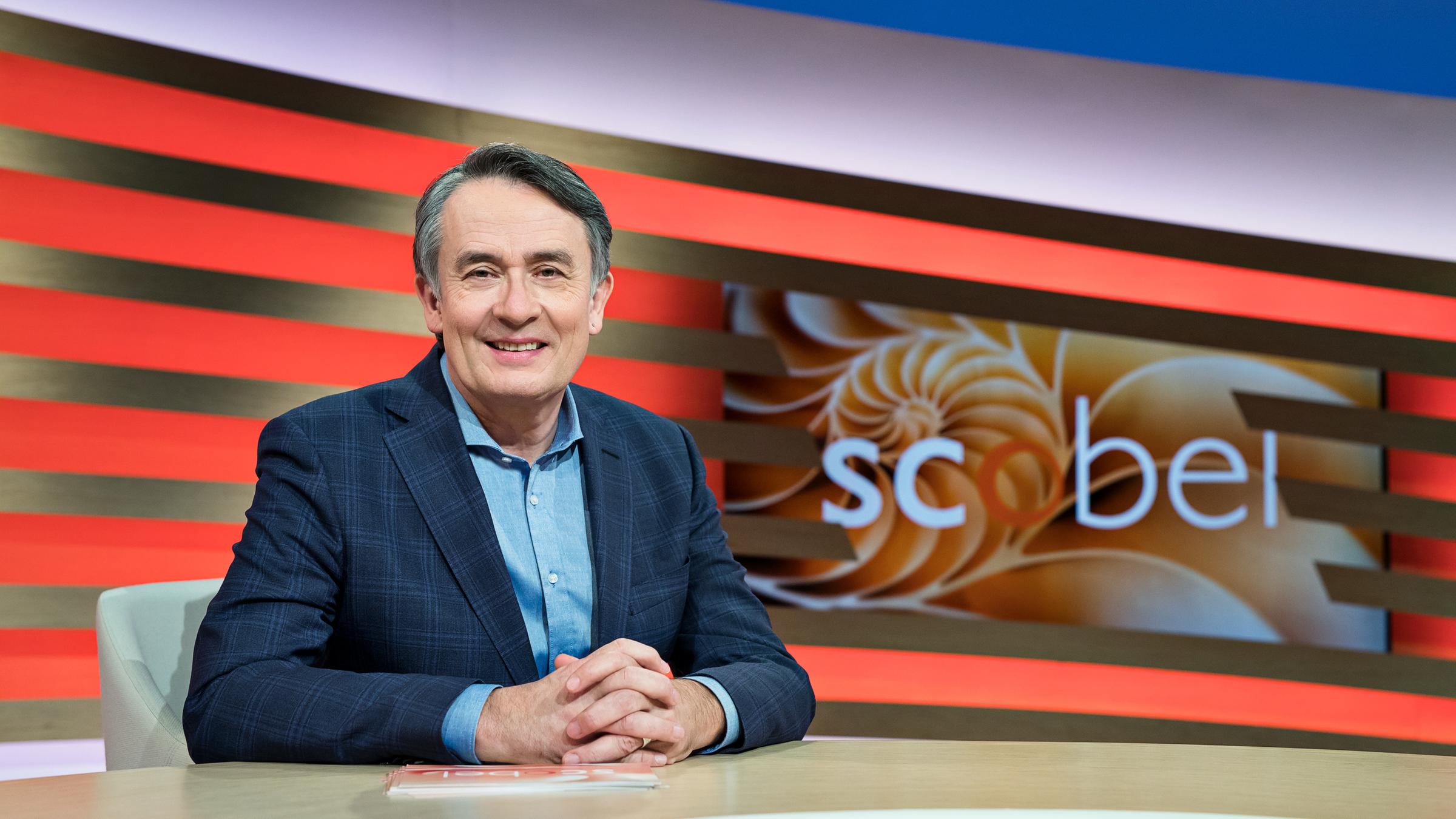 "scobel": Gert Scobel sitzend am Tisch in Studio-Deko