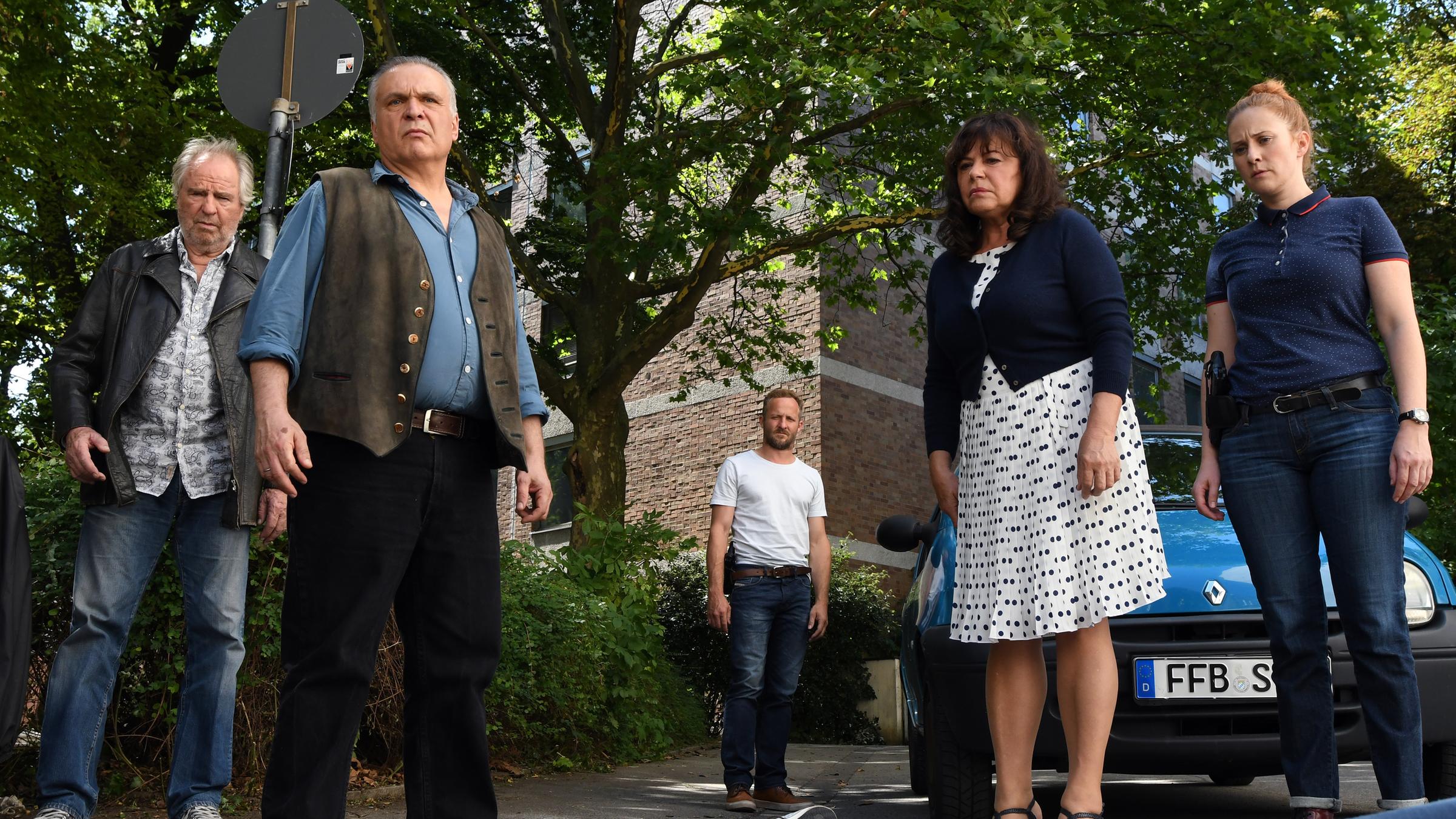 Schwarzach 23 und der Schädel des Saatans": Franz Germinger Senior (Friedrich von Thun), Karl Obermaier (Jockel Tschiersch), Franz Germinger Junior (Maximilian Brückner), Erika Germinger (Gundi Ellert) und Anna Germinger (Marlene Morreis) stehen vor einem Auto und betrachten eine Person, die vor ihnen am Boden liegt.