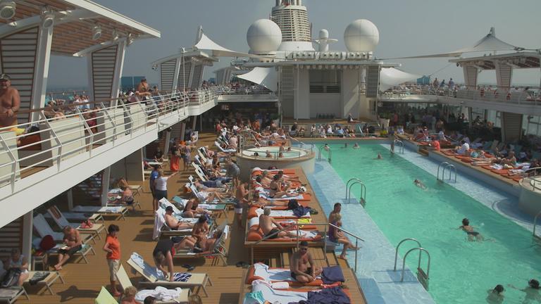 "ZDFzeit: AIDA, TUI & Co. - Der Kreuzfahrt-Check": Viele Menschen liegen in Badebekleidung auf Liegen auf dem Pooldeck eines Kreuzfahrtschiffes. Einige schwimmen.