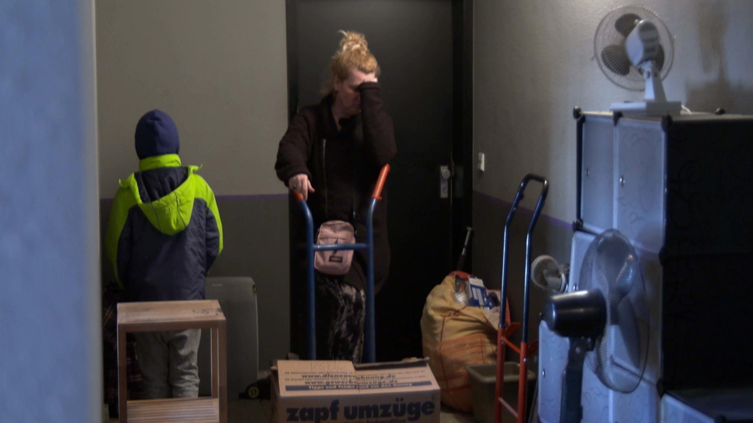 "Frontal 21-Dokumentation - Teurer Wohnen": Eine Frau steht in einem Hausflur und stützt ihren Kopf auf einem Arm ab. Vor ihr ist ein Umzugskarton zu sehen. Ein kleiner Junge, der nur von hinten zu sehen ist, steht neben der Frau.
