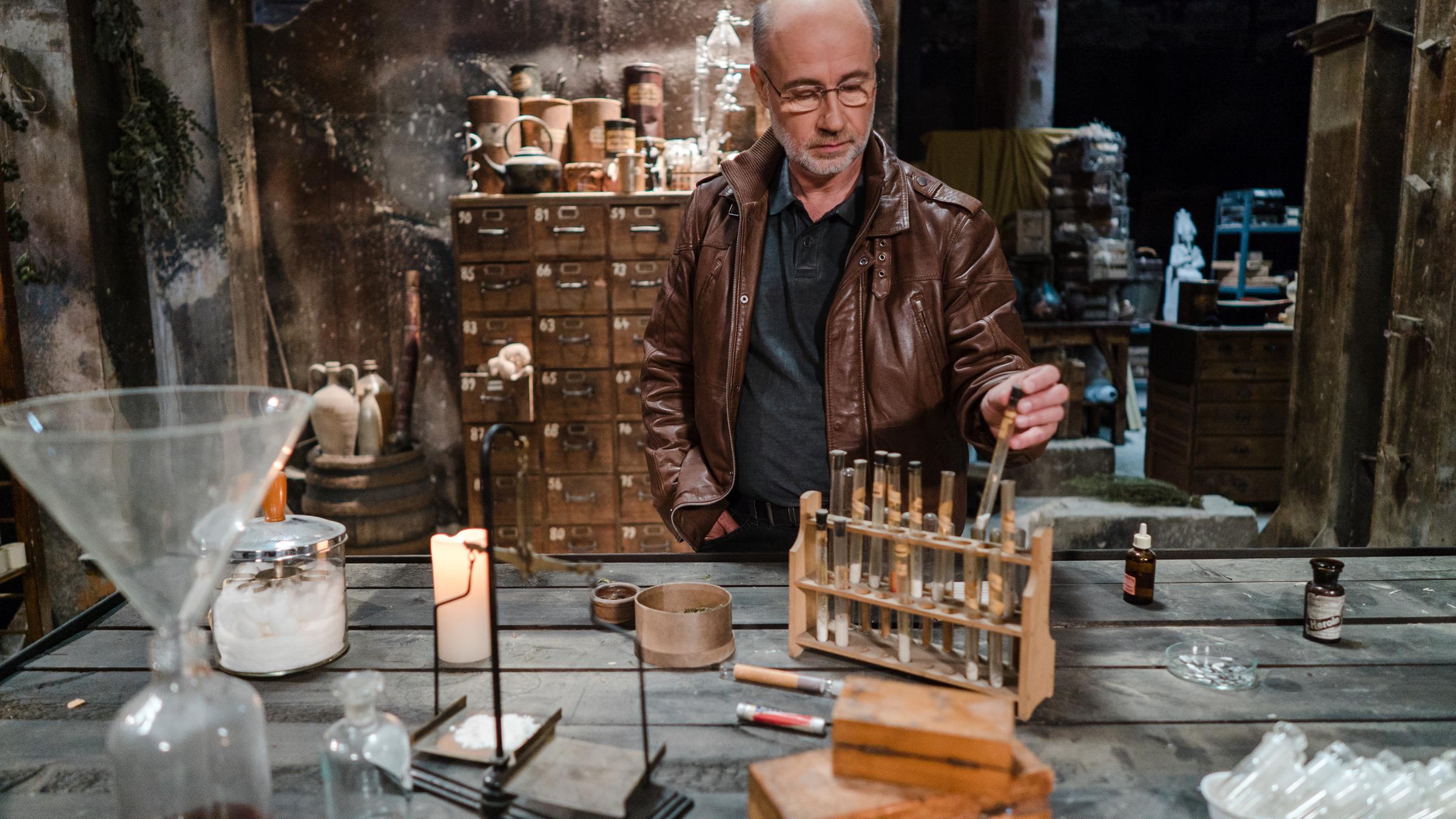 "Terra X: Drogen - Eine Weltgeschichte - Zwischen Medizin und Missbrauch": Moderator Harald Lesch steht an einem Tisch in einer alten Apotheke und zieht ein Reagenzglas aus einer Holzvorrichtung, in der noch weitere gefüllte Reagenzgläser stehen und andere Gegenstände zur Herstellung von Medizin.