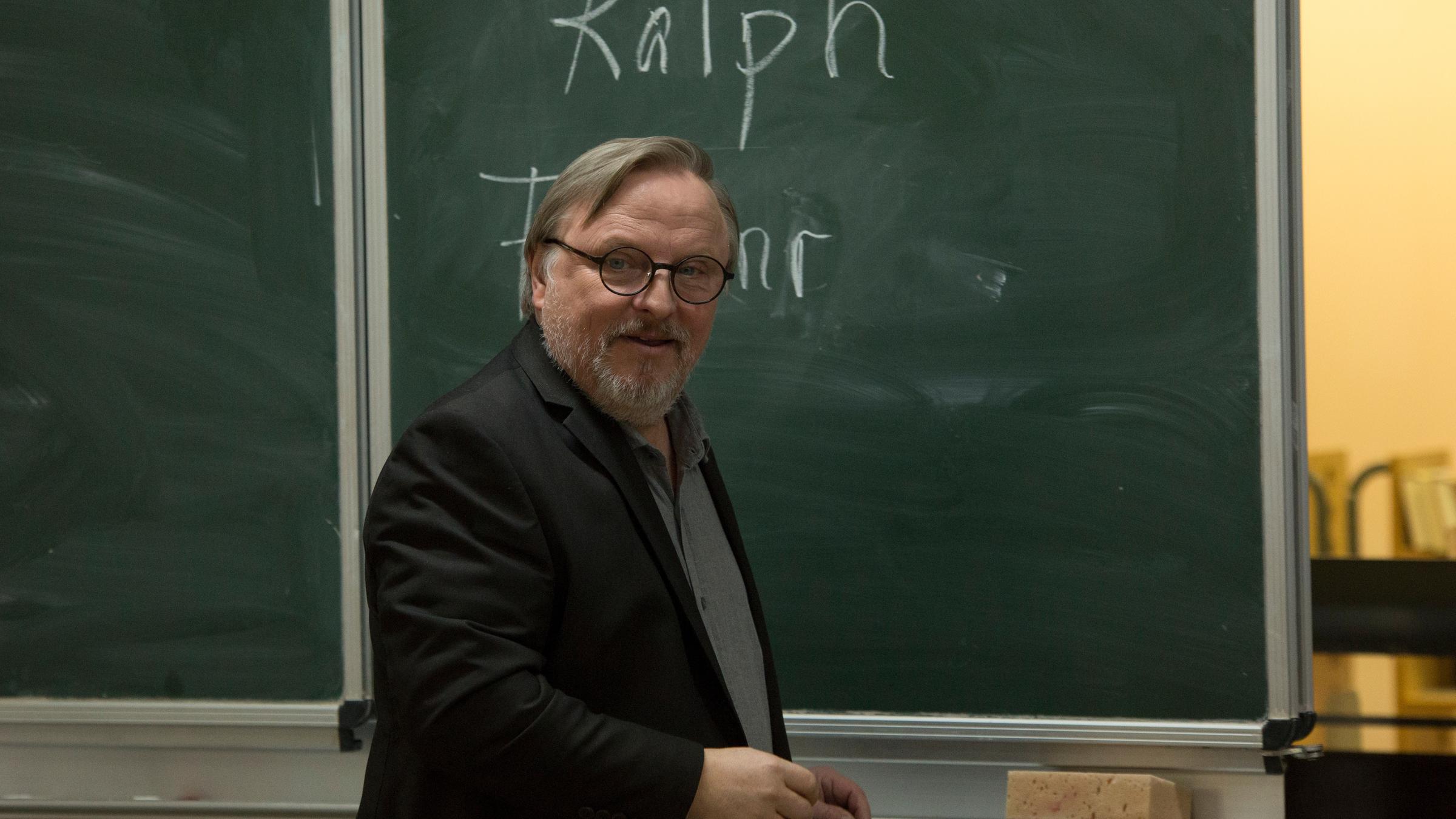 Ralph Friesner (Axel Prahl) steht seitlich zu einer Schultafel, sein Blick ist frontal gerichtet. Auf der Tafel ist sein Name geschrieben, wobei nur der Vornahme vollständig zu sehen ist.