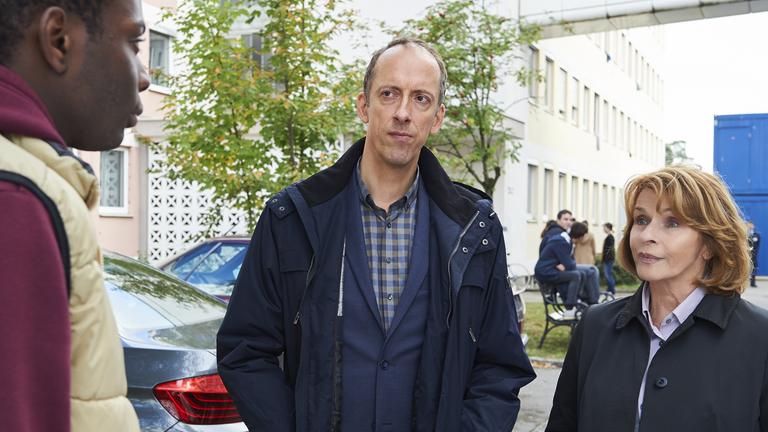 "Unter Verdacht - Die Guten und die Bösen": Chandu (Bortey Wendler) steht auf der Straße vor der Flüchtlingsunterkunft und sieht zu André Langner (Rudolf Krause) und Dr. Eva Maria Prohacek (Senta Berger), die vor ihm stehen und ihn anschauen.