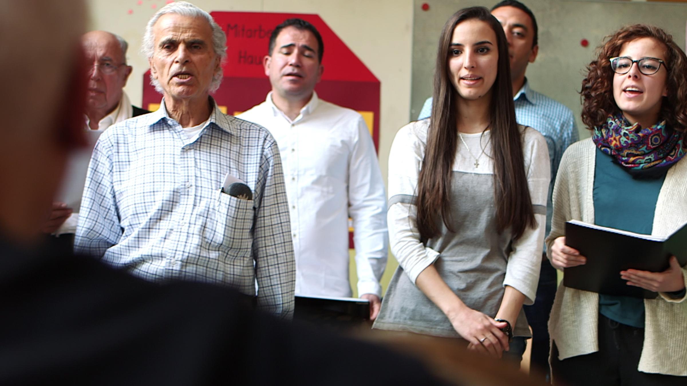 "plan b: Die Multi-Kulti-Macher - Wie Integration gelingen kann": Ältere und jüngere Frauen und Männer stehen zusammen und singen. Eine Frau hält eine Mappe in der Hand.