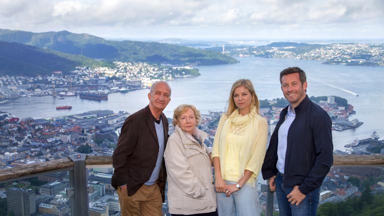 "Kreuzfahrt ins Glück - Hochzeitsreise nach Norwegen": Joachim Reiter (Philip Sonntag), Marlene Reiter (Christine Schorn), Stefanie von Erlen (Hilde Dalik) und Tom (Jan Hartmann) stehen auf einer Aussichtsplattform nebeneinander und lächeln in die Kamera. Im Hintergrund sieht man eine Stadt, die direkt an der See liegt.