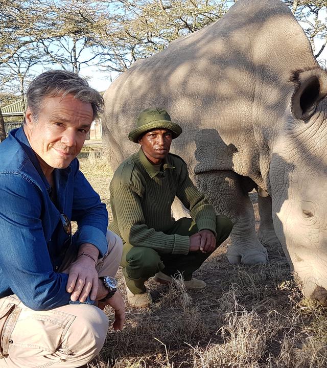 "Hannes Jaenicke: Im Einsatz für Nashörner": Hannes Jaenicke und ein Ranger knien im Freien neben dem Nashorn Sudan.