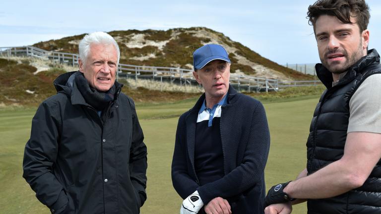 "Nord Nord Mord - Clüver und der leise Tod": Theo Clüver (Robert Atzorn) steht mit dem Immobilienmakler Bendix Böckelmann (Wilfried Hochholdinger) und dessen Assistenten Severin Laiser (Florian Jahr) auf dem Grün eines Golfplatzes in den Dünen von Sylt. Alle schauen in die gleiche Richtung.