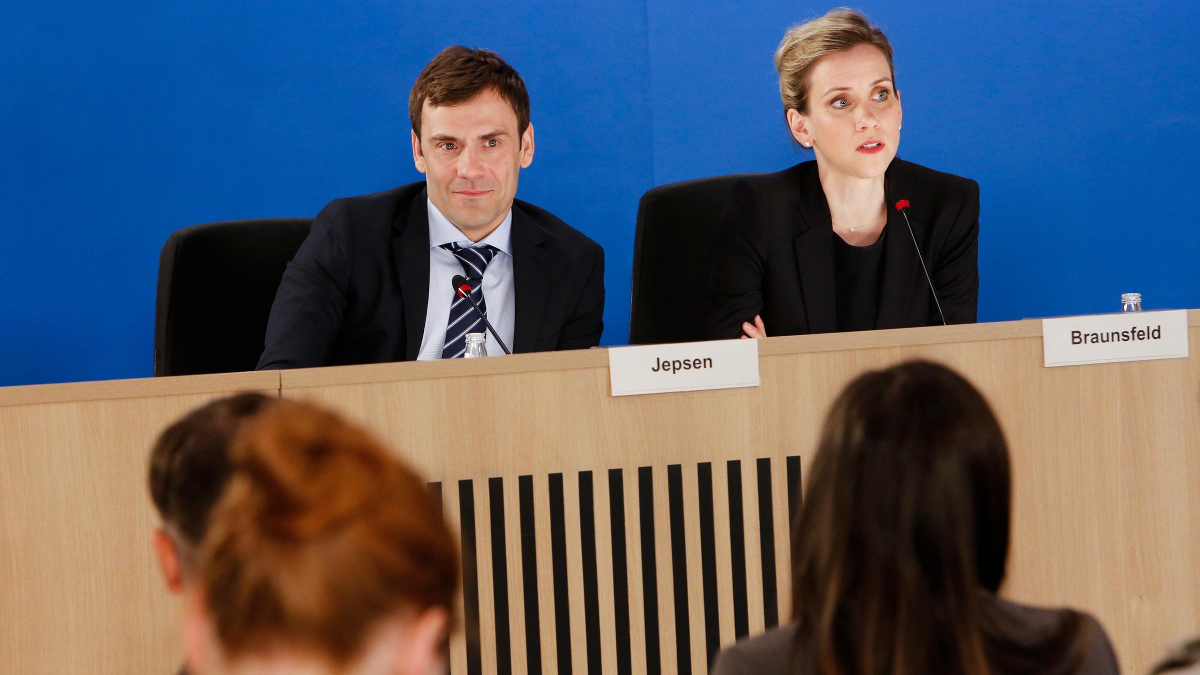 "Danke Deutschland!": Der Regierungssprecher Ludger Jepsen (Daniel Wiemer) sitzt neben der Pressesprecherin Gerda Braunsfeld (Lena Dörrie) an einem langen Pult. Vor ihnen sitzen Journalisten, welche nur schemenhaft zu erkennen sind.