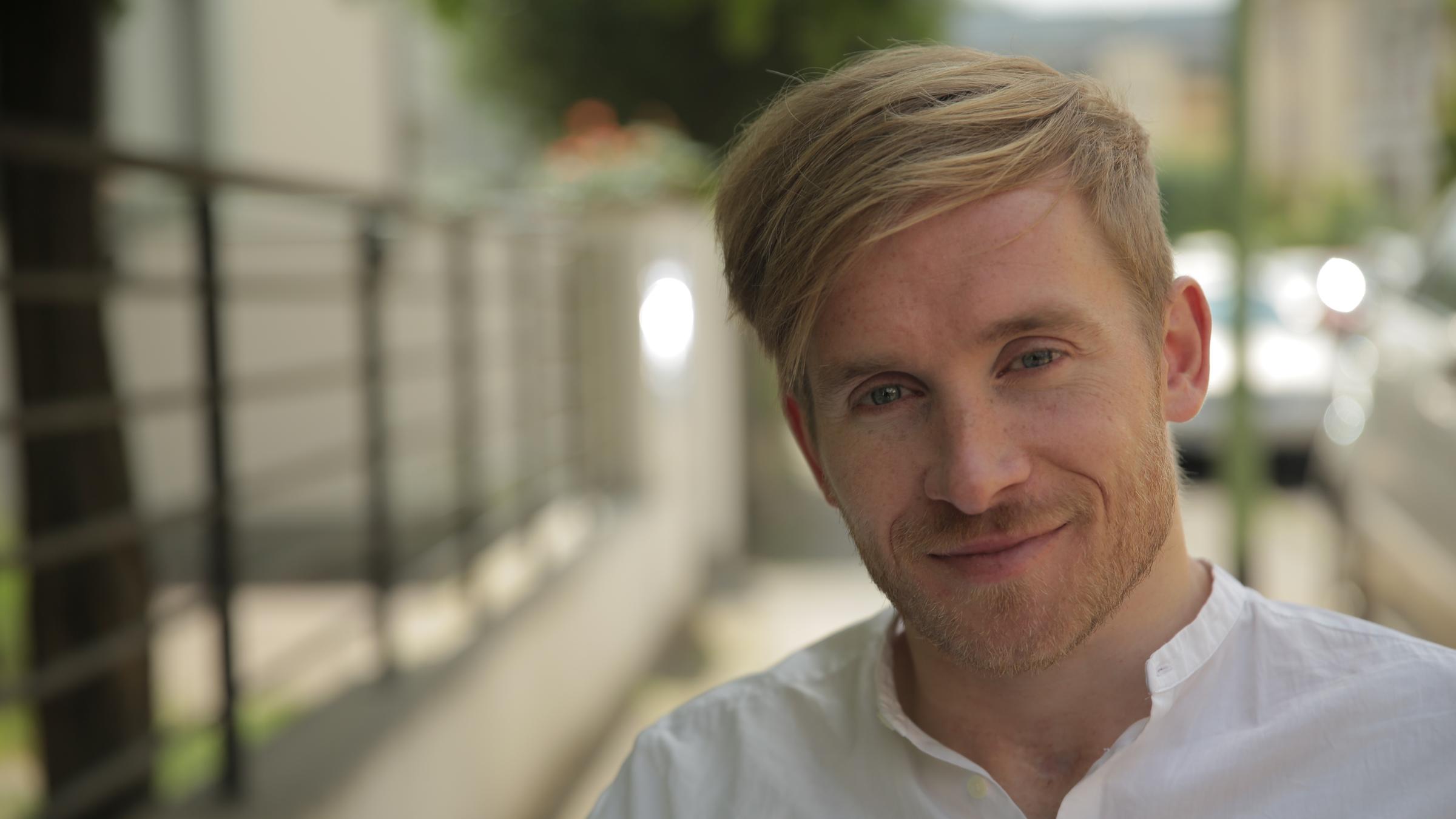 "37°: Vorwärts ins Leben - Auf dem Weg mit Samuel Koch": Porträtaufnahme von Samuel Koch im weißen Hemd. Er schaut in die Kamera. Im Hintergrund unscharf ein Bürgersteig mit parkenden Autos und Gartenzaun.