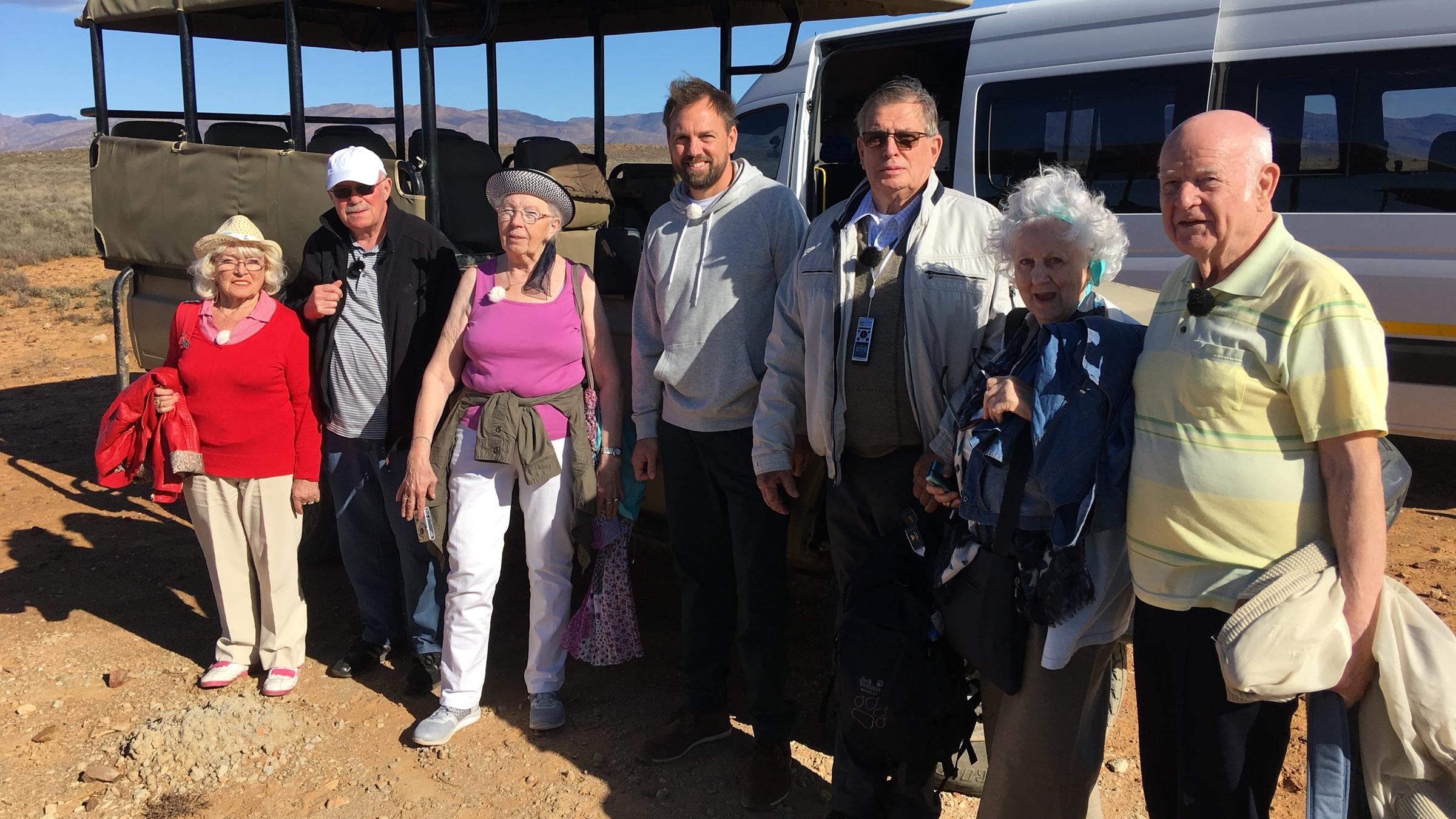Mit 80 Jahren um die Welt": Die Reisegruppe, bestehend aus Erika, Bernd, Marianne, Lothar, Christina und Norbert, steht zusammen mit Steven Gätjen vor einem Shuttlebus in der südafrikanischen Steppe. Alle lächeln in die Kamera.