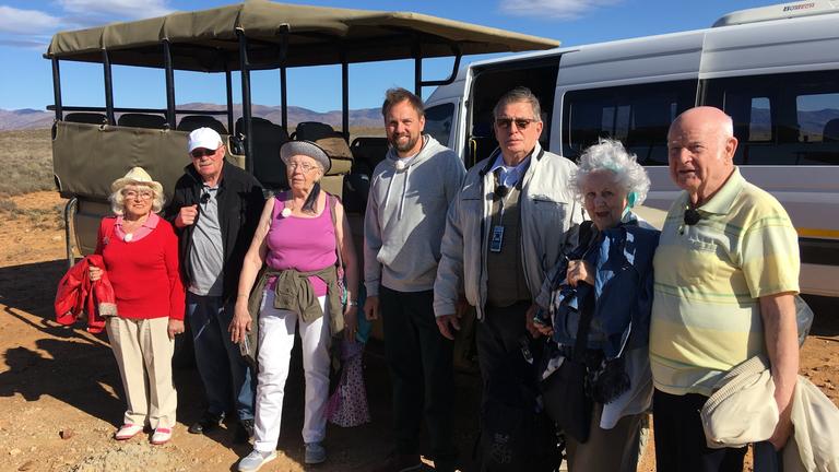 Mit 80 Jahren um die Welt": Die Reisegruppe, bestehend aus Erika, Bernd, Marianne, Lothar, Christina und Norbert, steht zusammen mit Steven Gätjen vor einem Shuttlebus in der südafrikanischen Steppe. Alle lächeln in die Kamera.
