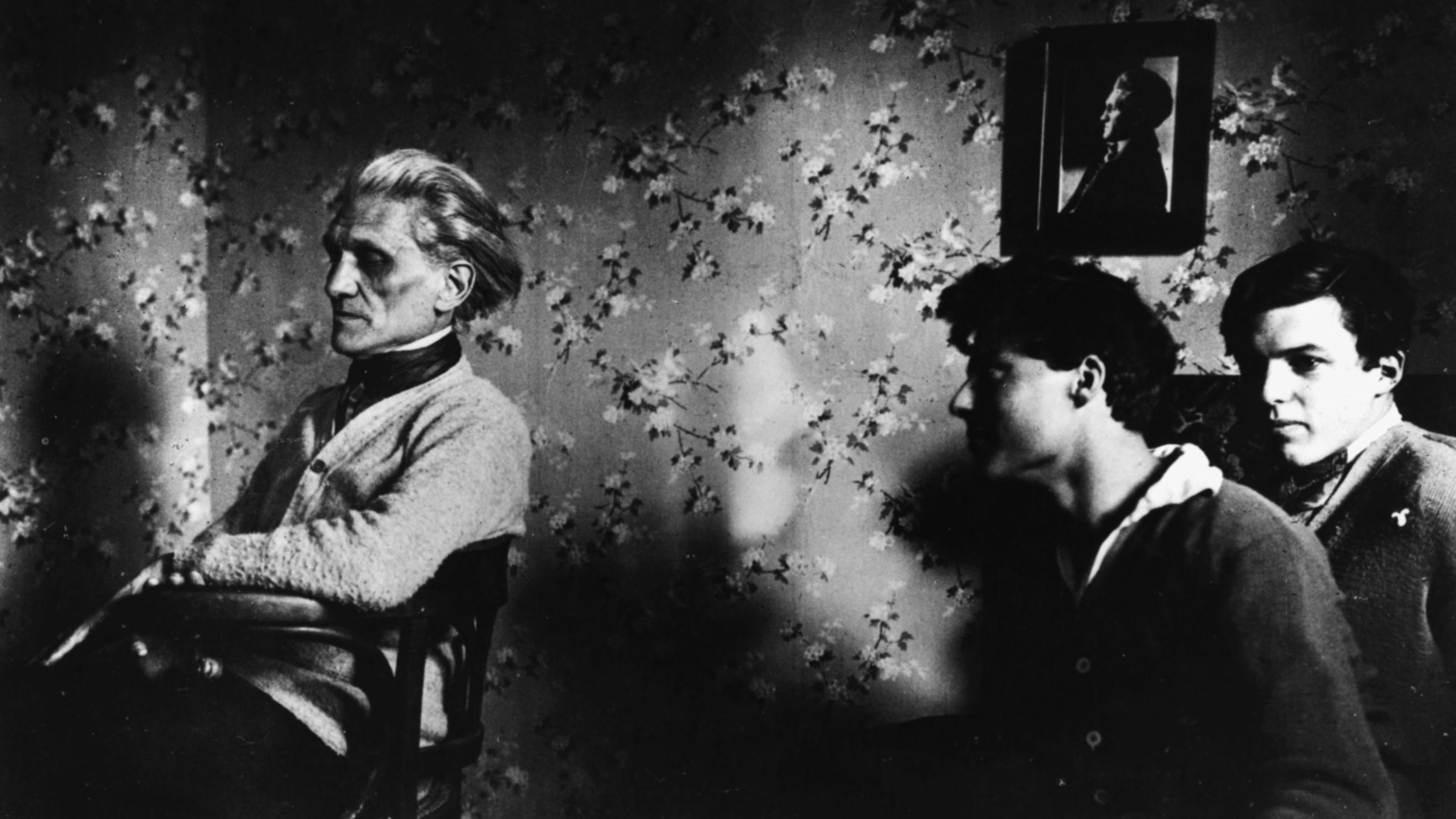 "Stefan George: Das geheime Deutschland": George sitzt auf einem Stuhl und blickt nach links aus dem Bild. Die beiden sehr jungen Stauffenbergs sitzen in seinem Rücken, blicken zu ihm auf.