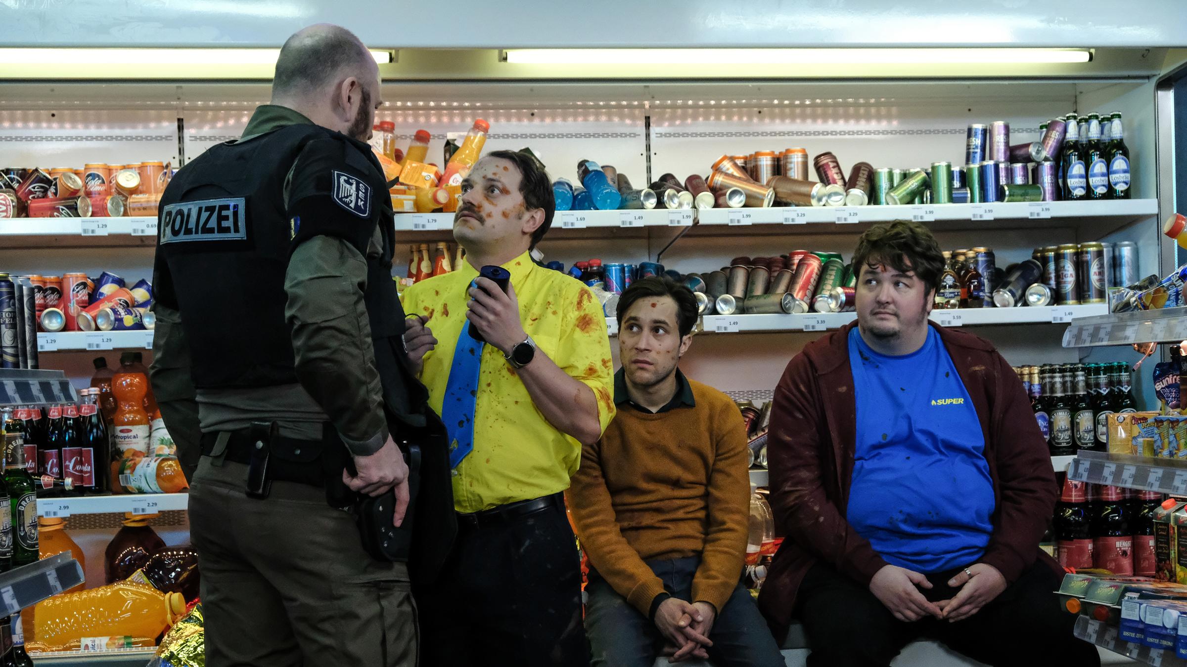 "Tanken - Mehr als Super: Kundenterror": Der Einsatzleiter (Alexander Grünberg) steht in der Tankstelle Georg Bergstedt (Stefan Haschke) mit der Hand an der Waffe gegenüber. Daniel (Ludwig Trepte) und Olaf (Daniel Zillmann) sitzen auf der Theke vor den Regalen und beobachten gespannt die Situation. Die drei Tankstellen-Mitarbeiter sind mit einer roten Flüssigkeit besudelt.