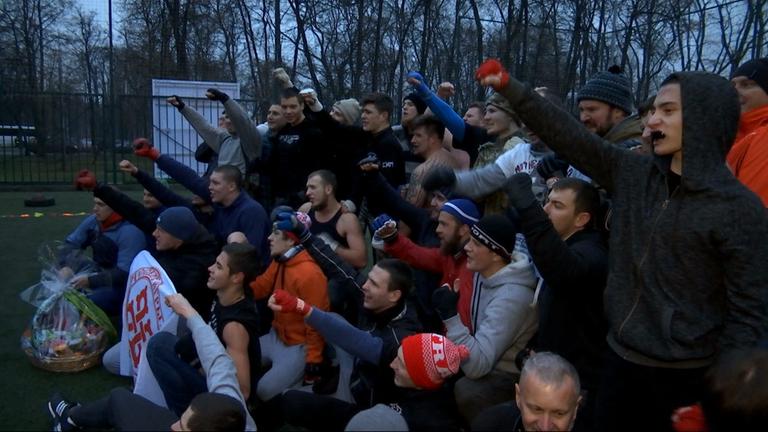 "Kampfbereit: Russlands Hooligans - Fußball, Randale und Politik": Mehrere männliche, russische Hooligans stehen, sitzen oder hocken in einer Gruppe zusammen, schauen alle in die gleiche Richtung und einige von ihnen halten einen Arm nach oben.