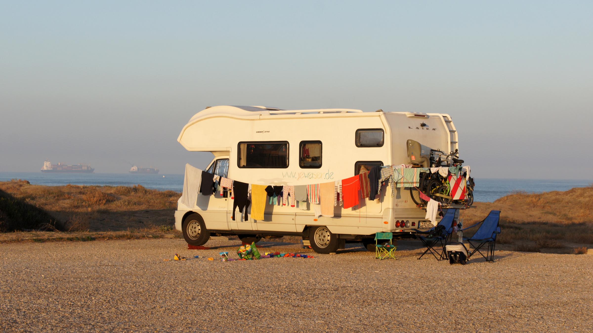 "37°: Mein Haus zieht mit - Abenteuer Hausboot, Camper, Tiny House": Der Camper steht allein auf einem Parkplatz, im Hintergrund sieht man das Meer. Entlang des Campers ist eine Wäscheleine gespannt, an der bunte Wäschestücke hängen.