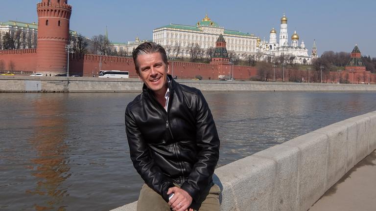 "Markus Lanz - Russland!": Markus Lanz sitzt auf einer Ufermauer und lacht in die Kamera. Im Hintergrund ist der Kreml in Moskau zu sehen.