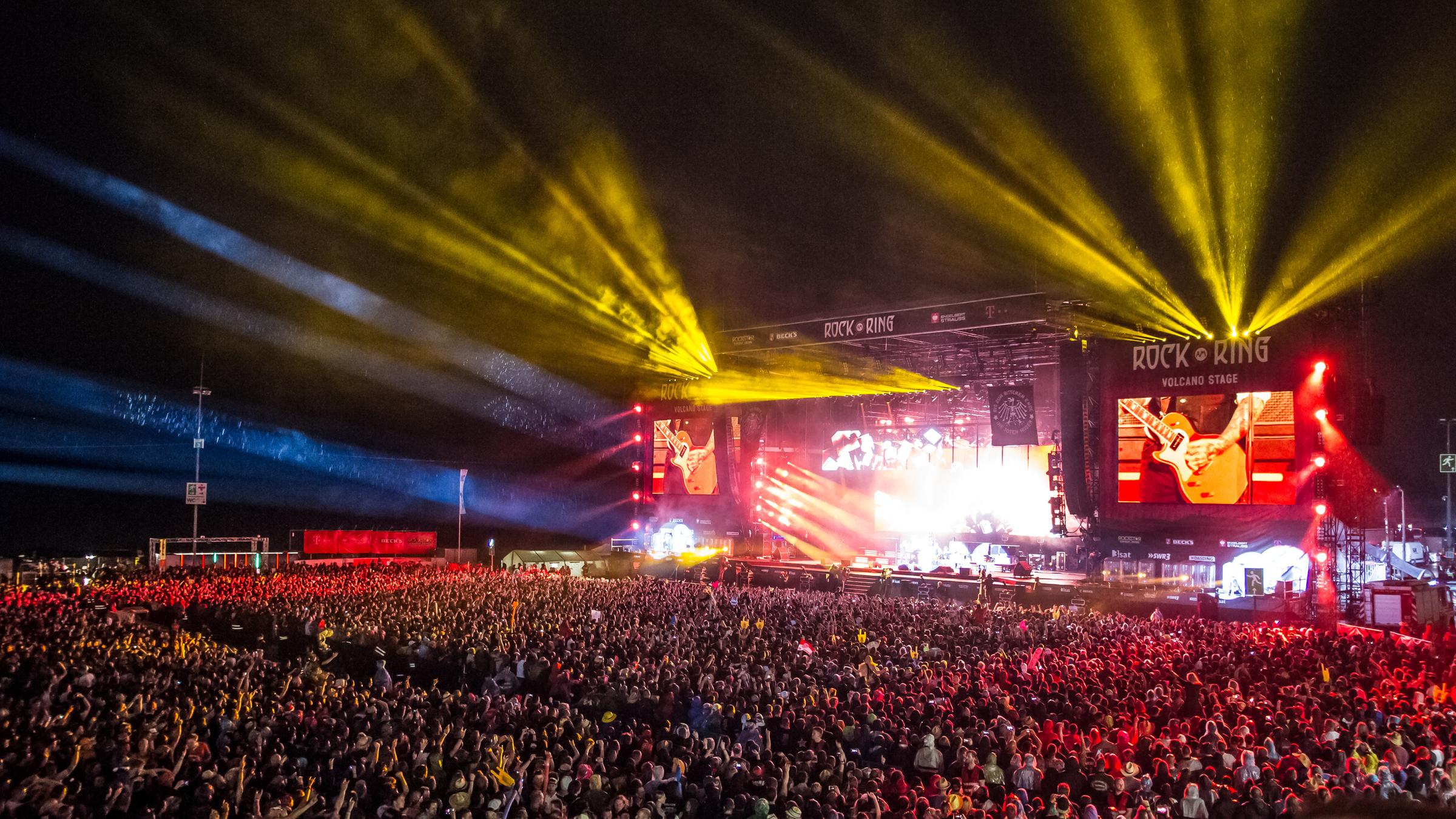 "Rock am Ring 2018": Rechts im Bild ist die Volcano Stage zu sehen, eine der beiden Hauptbühnen von „Rock am Ring“. Es ist Nacht, die Bühne ist beleuchtet. Davor stehen tausende von Menschen, man sieht die Absperrungen zwischendrin.
