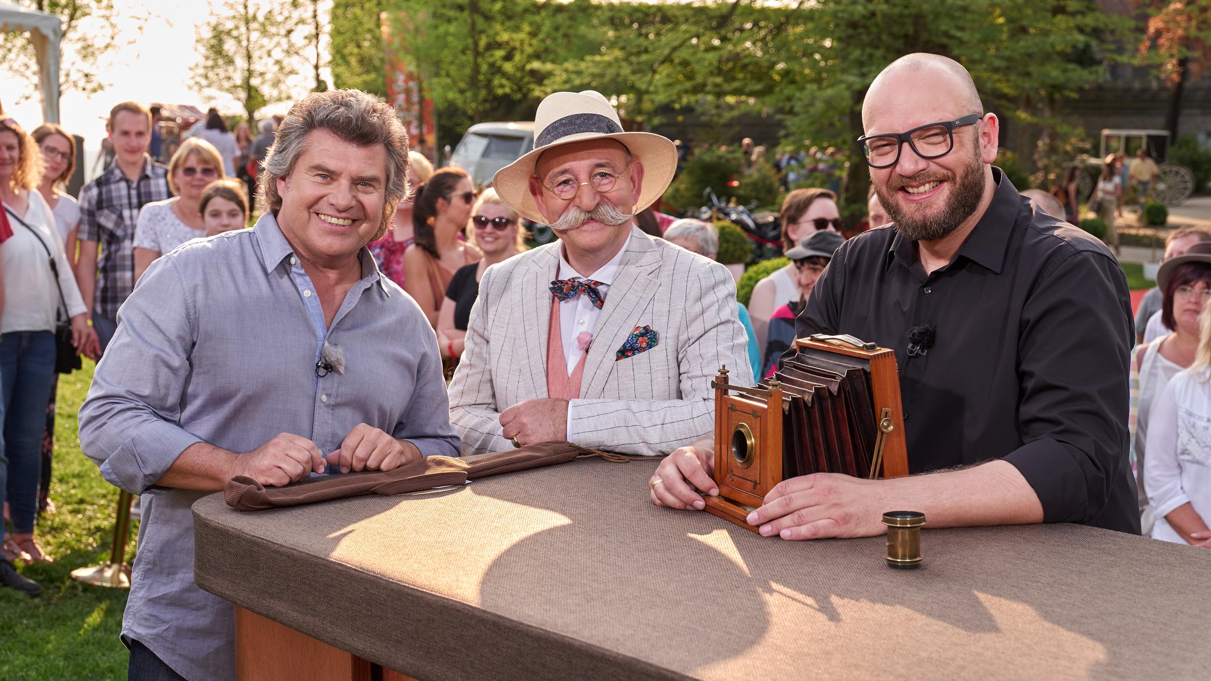"Bares für Rares - Deutschlands größte Trödelshow": Andy Borg, Horst Lichter und Sven Deutschmanek stehen hinter einem Tisch und lächeln. Vor Sven Deutschmanek steht das Objektiv einer alten Plattenkamera.