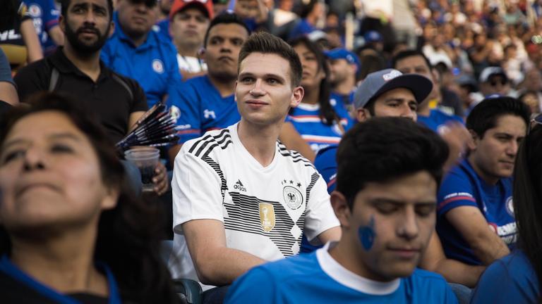 "Fußball-Fieber - Das WM-Magazin: Fabian Kösters Gruppengegner-Check": Fabian Köster sitzt im DFB-Trikot auf der Tribüne. Um ihn herum sitzen viele Fans in blauen Trikots.