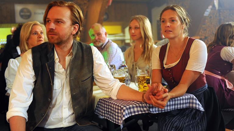 "Drachenjungfrau": Alma (Patricia Aulitzky) sitzt Händchen haltend mit Robert Meierhofer (Markus Hamele) im Wirtshaus an einer Bierzeltgarnitur. Beide schauen in die gleiche Richtung. Am Tisch sitzen weitere Gäste (Komparsen), auf dem Tisch stehen Biergläser.