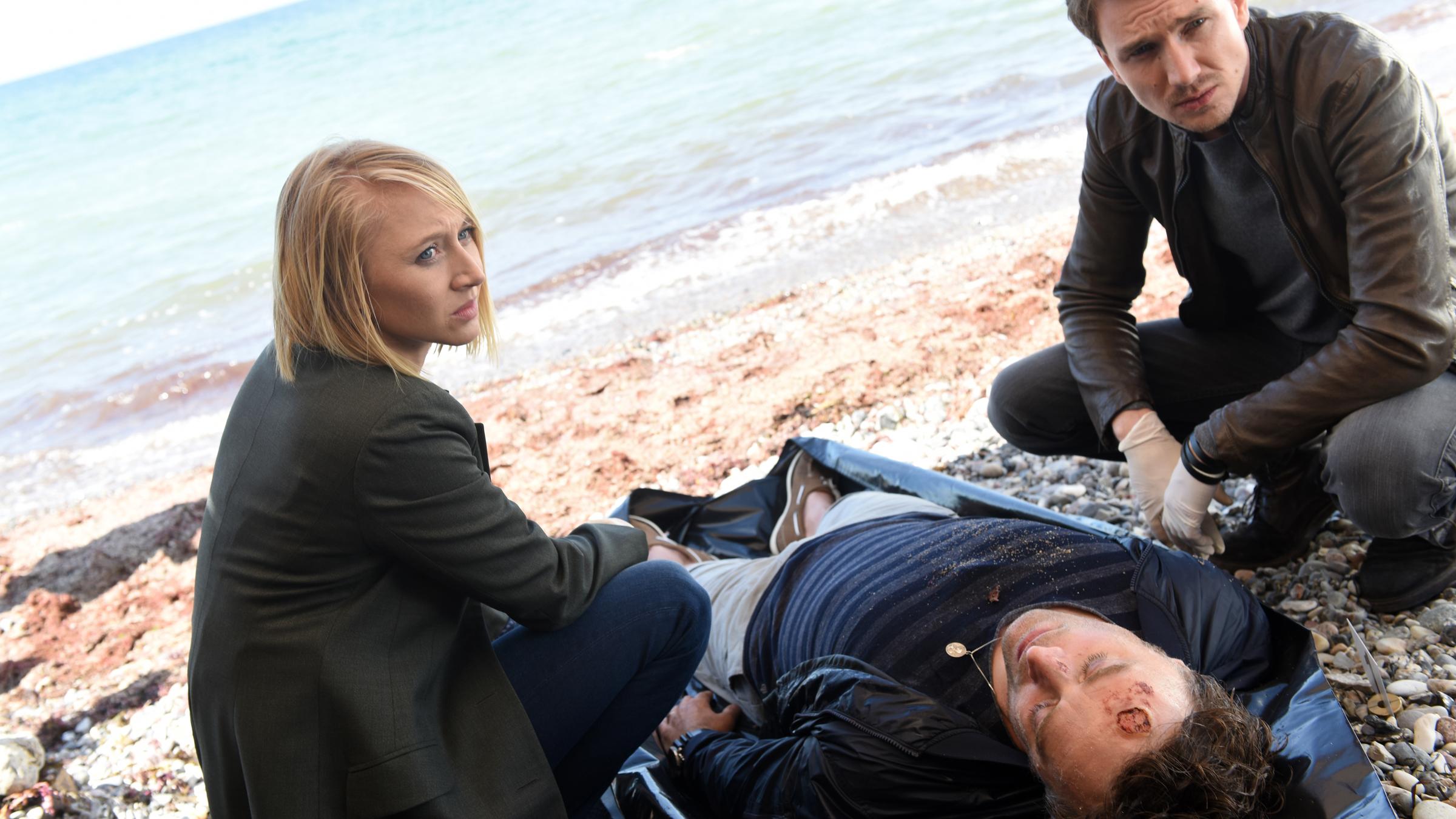 "Solo für Weiss - Es ist nicht vorbei": Nora Weiss (Anna Maria Mühe) und Simon Brandt  (Jan Krauter) sitzen neben der Leiche Patrick Landau (Eric Langner) am Strand.