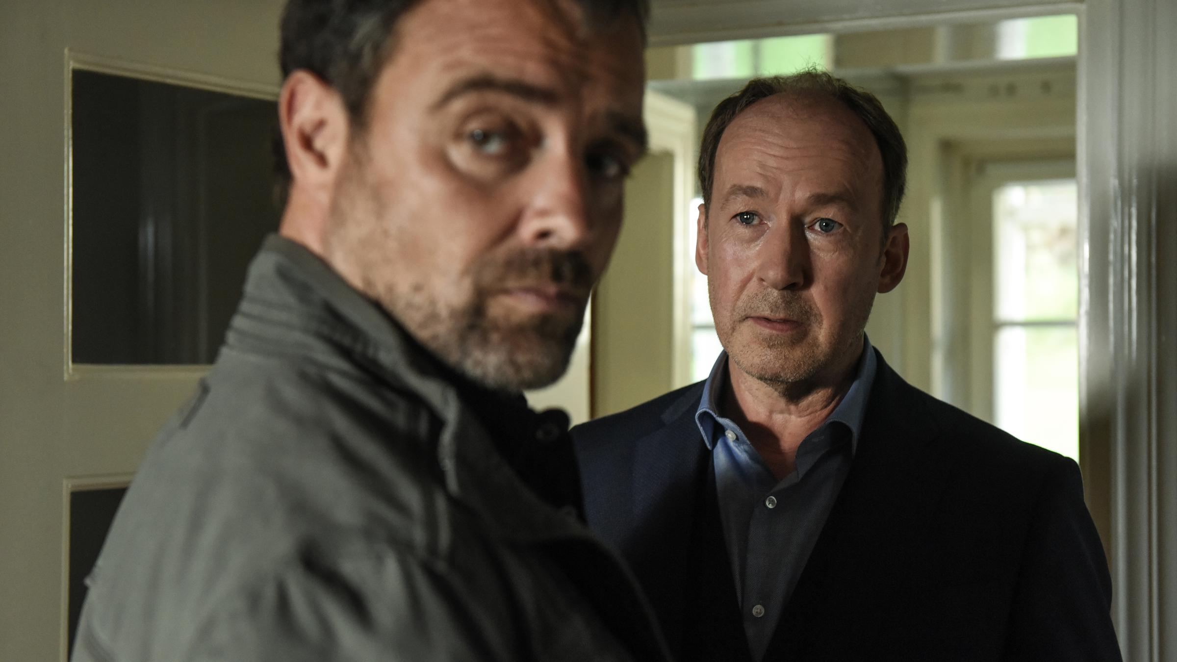 "Neben der Spur – Sag, es tut dir leid": Joe (Ulrich Noethen) steht in einer geöffneten Zimmertür und schaut Ruiz (Jürgen Maurer) an, der ihm gegenübersteht und nach hinten schaut.