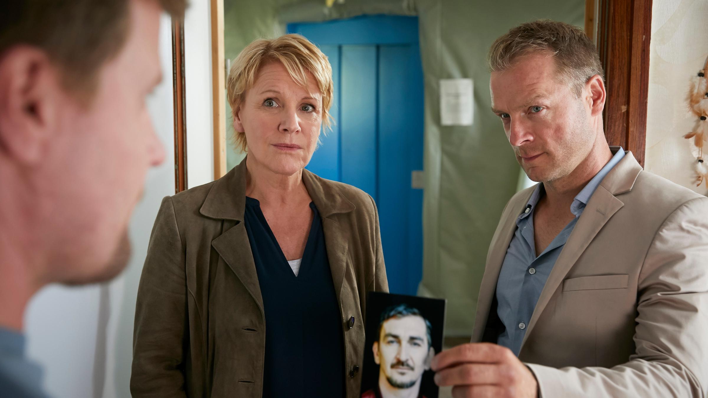 "Marie Brand und der Duft des Todes": Marie Brand (Mariele Millowitsch) und Jürgen Simmel (Hinnerk Schönemann) stehen Oliver Semper (Peter Moltzen) in einem Zimmer gegenüber. Simmel zeigt Semper ein Foto.