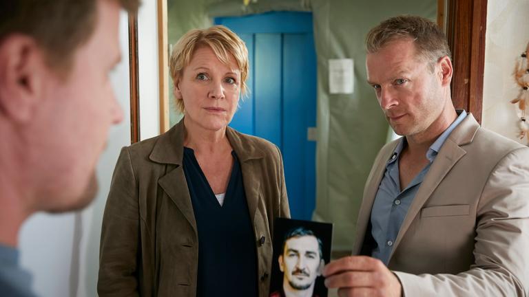 "Marie Brand und der Duft des Todes": Marie Brand (Mariele Millowitsch) und Jürgen Simmel (Hinnerk Schönemann) stehen Oliver Semper (Peter Moltzen) in einem Zimmer gegenüber. Simmel zeigt Semper ein Foto.