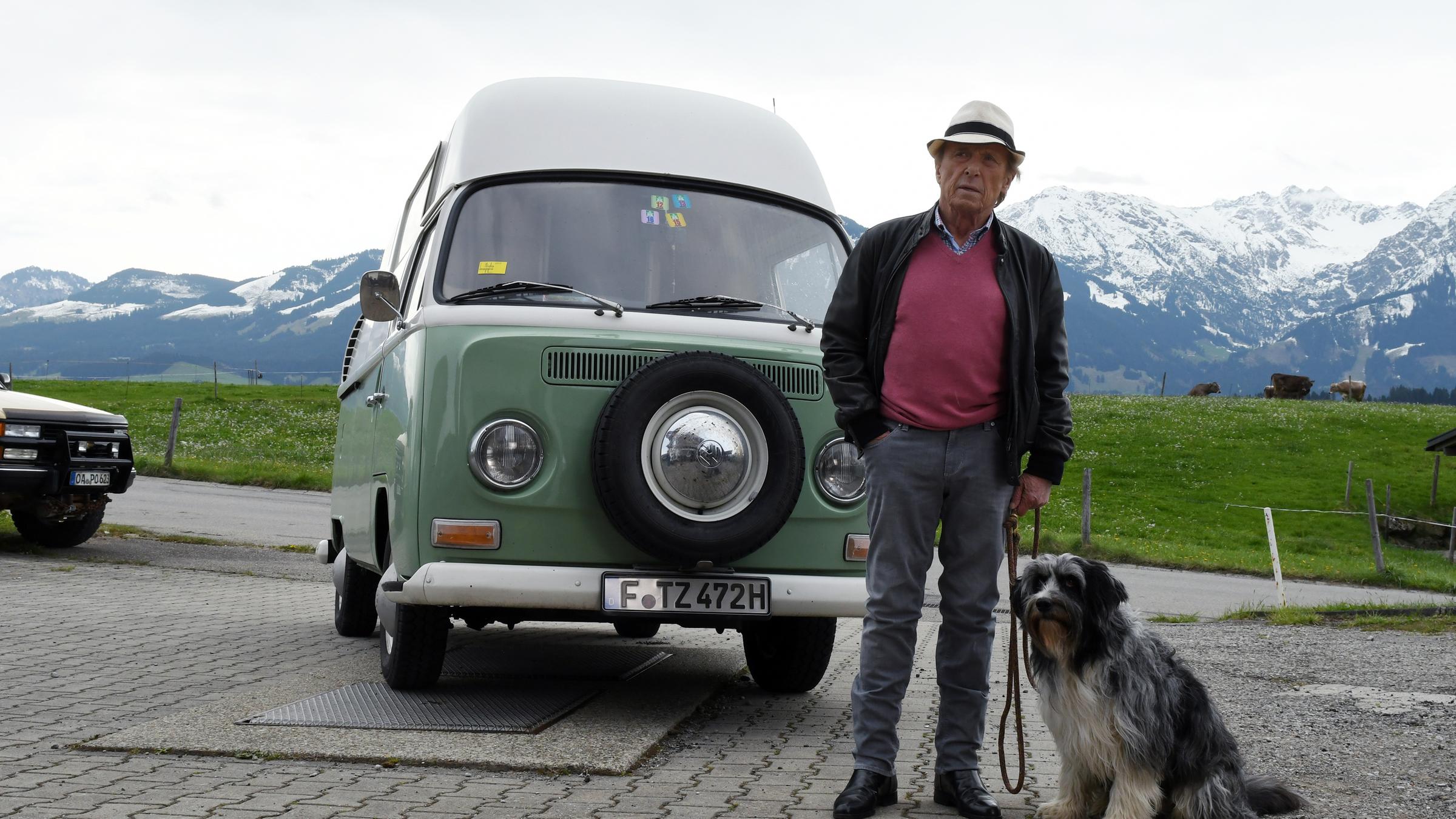 "Matula - Der Schatten des Berges": Matrula, sein Hund "Dr. Renz" und sein alter VW-Bus. Im Hintergrund eine Wiese und eine schneebedeckte Bergkette.