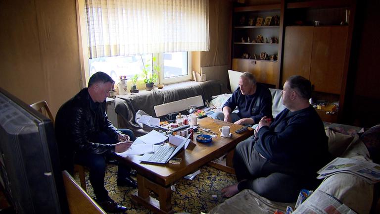 "ZDF.reportage: Geld oder Gefängnis?": Der Gerichtsvollzieher Frank Neuhaus sitzt in einem Wohnzimmer auf einem Stuhl und schaut auf die Unterlagen, die vor ihm auf dem Tisch liegen. Ihm gegenüber sitzen zwei Männer, die ins Leere blicken.