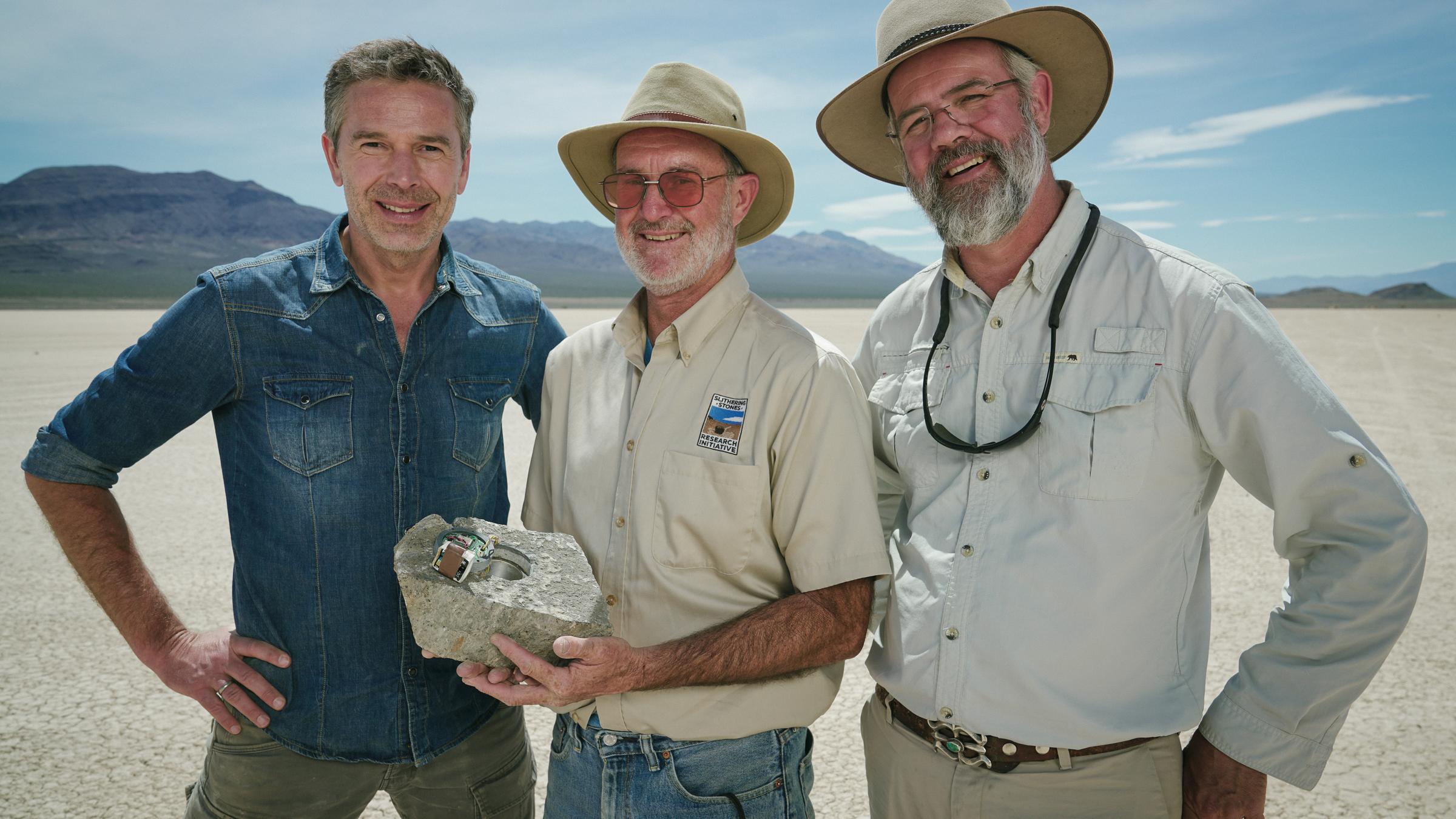 "Terra X: Rätselhafte Phänomene - Verschwundene Inseln und wandernde Steine": Dirk Steffens, Jim und Richard Norris stehen in einer Wüstenlandschaft nebeneinander und lachen in die Kamera. Jim hält einen präparierten Stein in  den Händen.