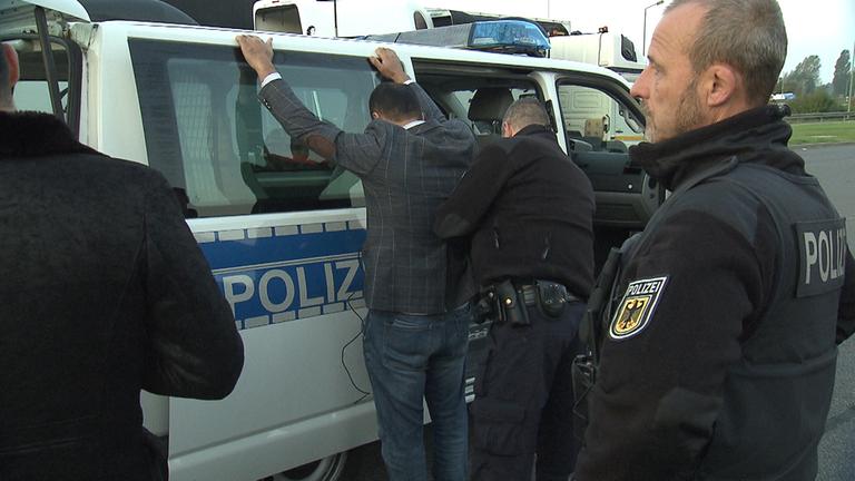 "ZDFzoom: Heimlich über die Grenze - Kampf gegen Schleuser": Polizisten überprüfen einen festgenommenen Mann, der mit erhobenen Händen an ein Polizeiauto gelehnt steht.