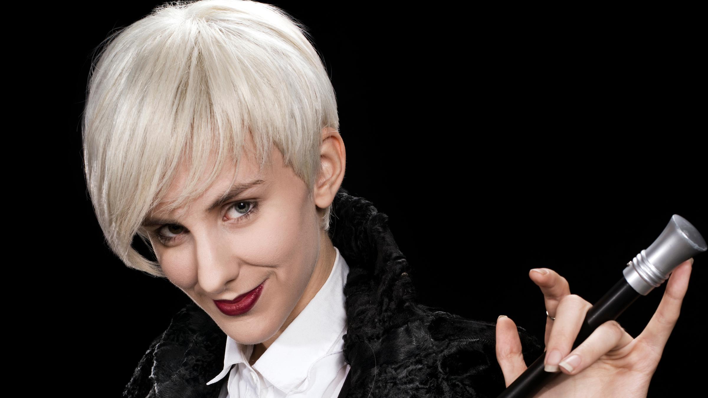 "Lisa Eckhart: Als ob Sie Besseres zu tun hätten": Lisa Eckhart hat weiße Haare und trägt einen Kurzhaarschnitt. Sie trägt eine enge schwarze Hose und eine weiße Bluse, dazu eine schwarze Weste und eine schwarze Samtjacke. Sie hält in ihren Händen einen schwarzen Stock mit silbernem Knauf.