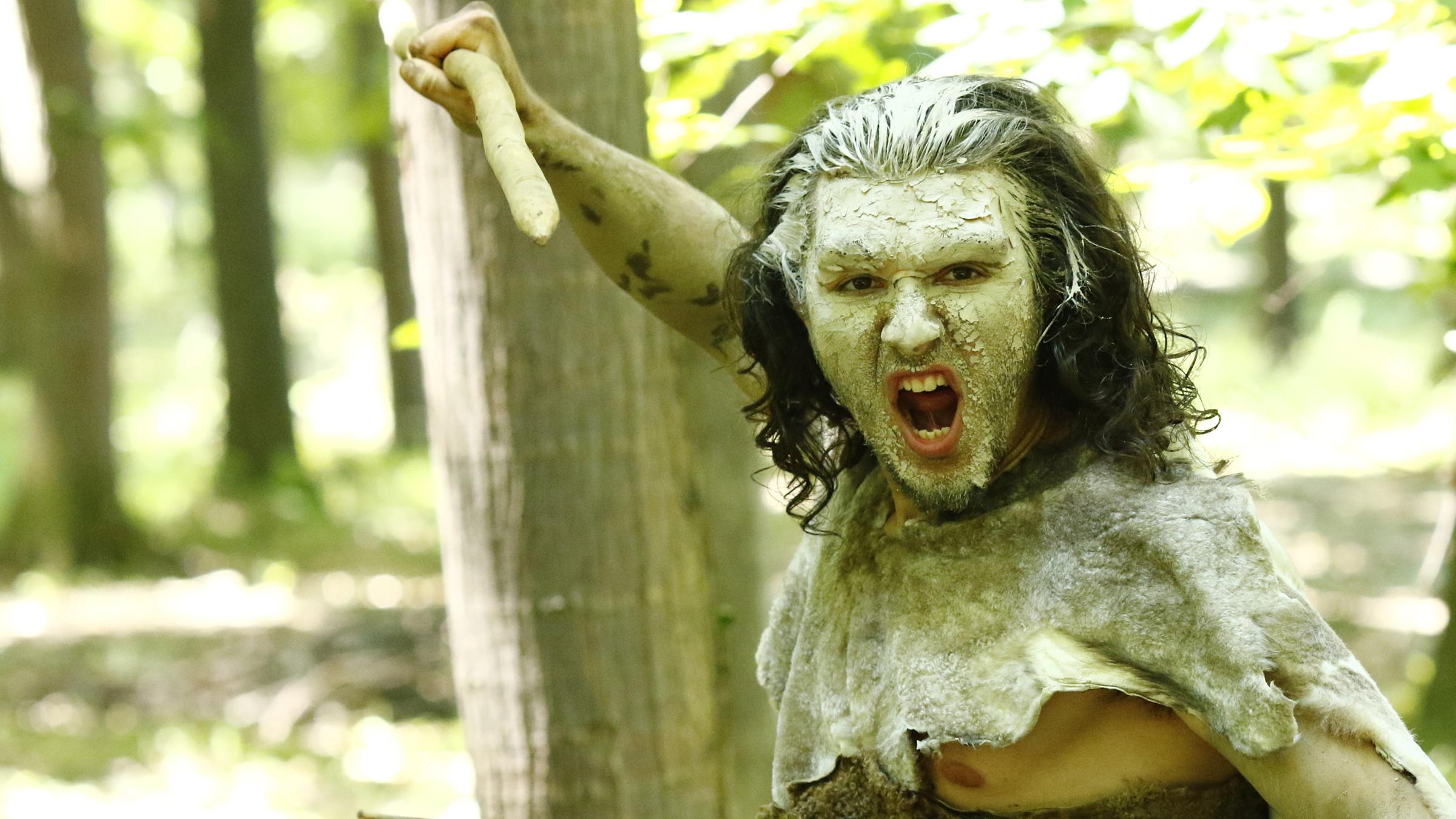 Ein in Felle gekleideter und im Gesicht weiß bemalter Steinzeitmensch in Kampfpose mit einem Holzspeer in jeder Hand.