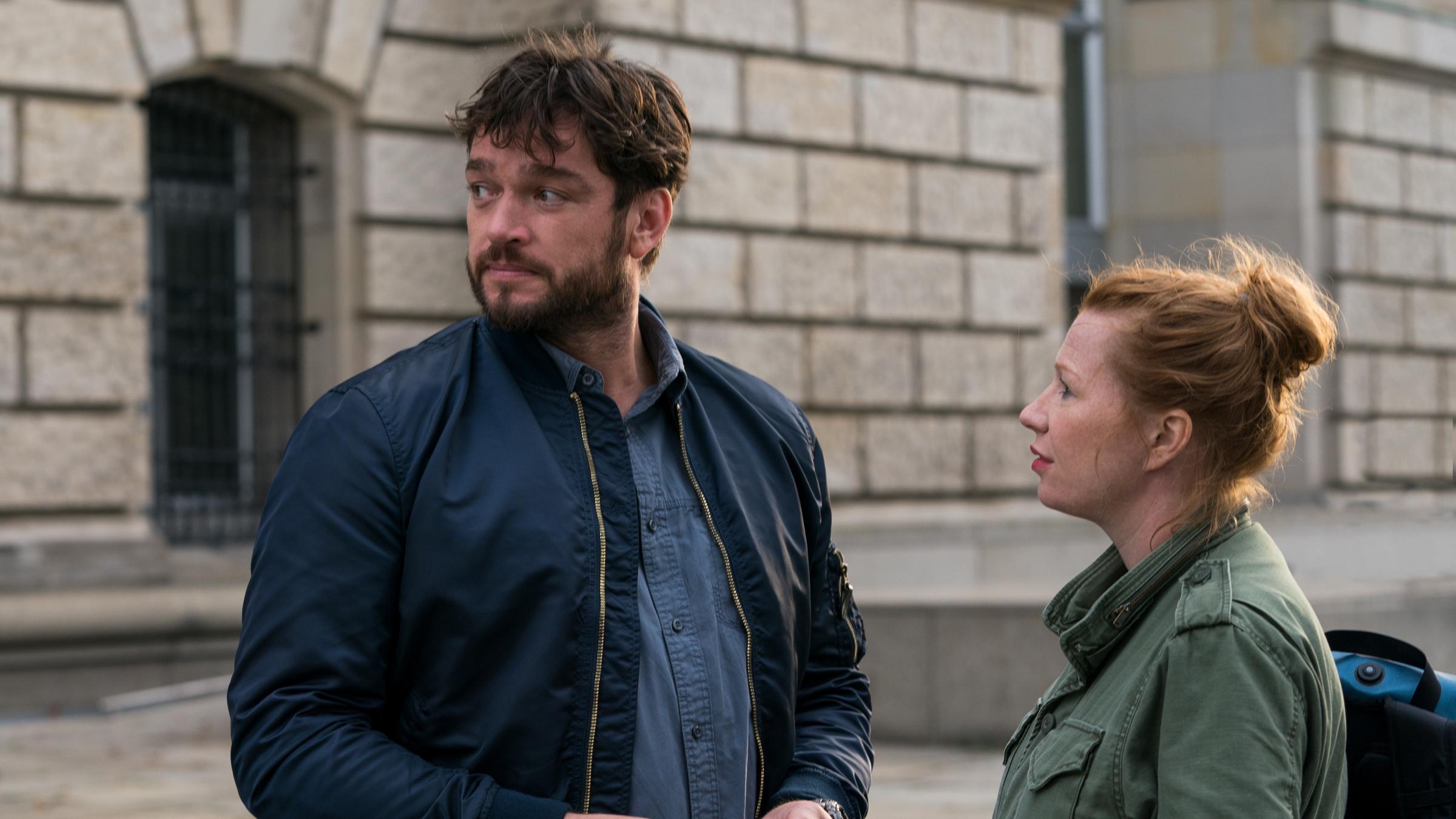 "Dengler - Fremde Wasser": Olga Illiescu (Birgit Minichmayr) und Georg Dengler (Ronald Zehrfeld) stehen zusammen auf einer Straße. Olga schaut Dengler ins Gesicht, während dieser über die rechte Schulter schaut und jemanden beobachtet.