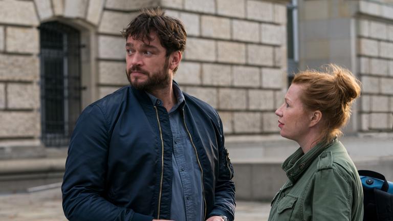 "Dengler - Fremde Wasser": Olga Illiescu (Birgit Minichmayr) und Georg Dengler (Ronald Zehrfeld) stehen zusammen auf einer Straße. Olga schaut Dengler ins Gesicht, während dieser über die rechte Schulter schaut und jemanden beobachtet.
