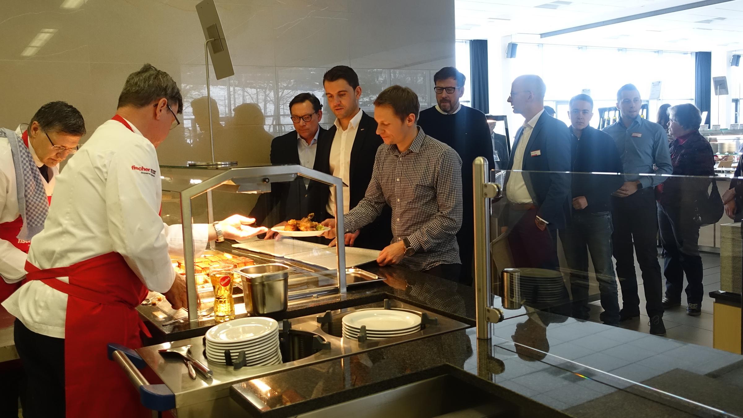 plan b: Mahlzeit! Köstliche Kantinenküche": Zwei Köche geben in der Kantine Essen aus, es hat sich eine Schlange wartender Arbeitnehmer gebildet.