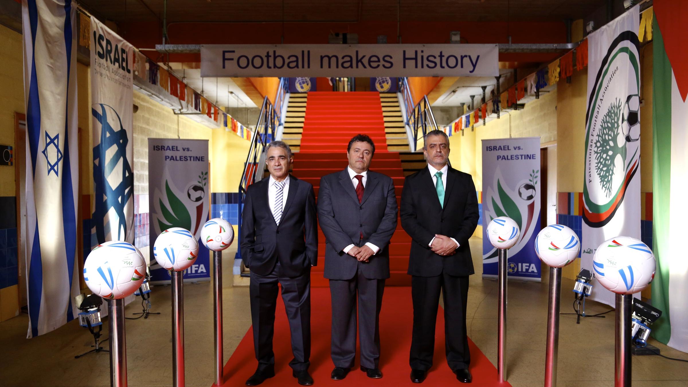 "Der 90-Minuten-Krieg": Der portugiesische Stadion-Manager Gomes (Alexander Barata) steht mit den Verbandsfunktionären Ozon aus Israel (Moshe Ivgy) und Barguti aus Palästina (Norman Issa) auf dem roten Teppich vor einer Treppe im Fußballstadion.