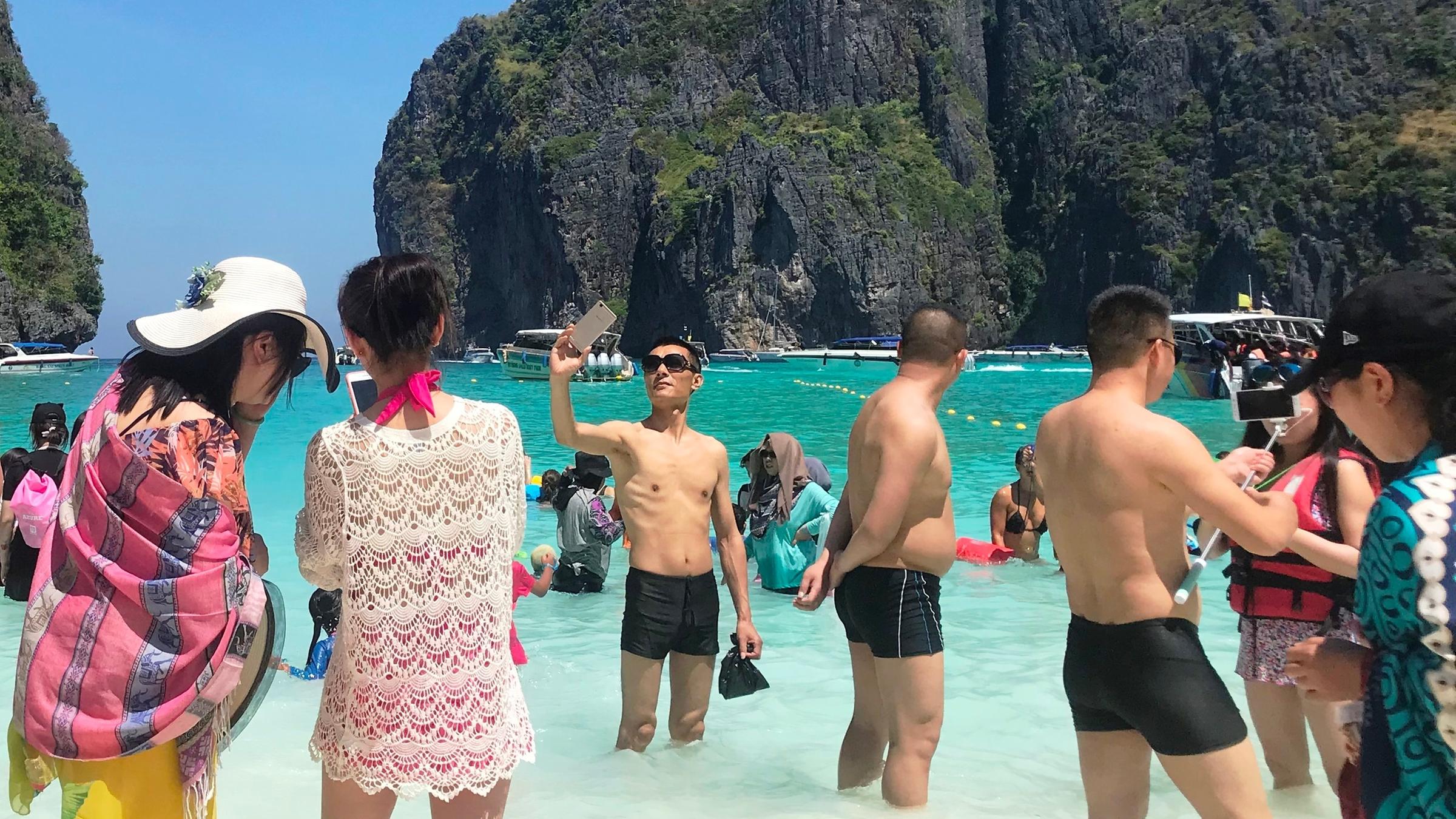 "Sommer, Sonne, Urlaubsglück?": Mehrere Personen stehen im und am Strand der Maya-Bucht in Thailand. Viele von ihnen machen Selfies. Im Hintergrund sind Felsen zu sehen.