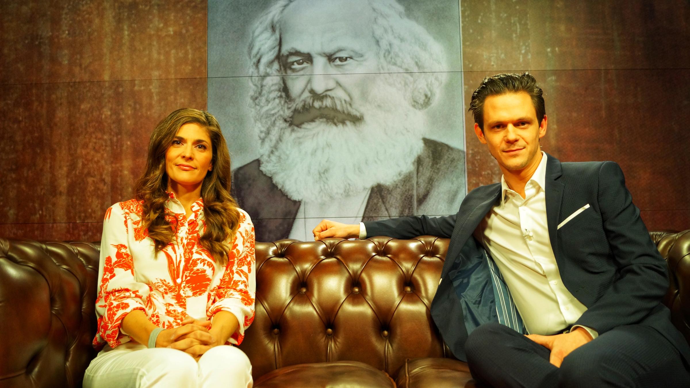 "aspekte - Deutsche Revolutionen": Katty Salié und Jo Schück sitzen auf einem Ledersofa. Zwischen beiden hängt eine große schwarz-weiß Fotografie von Karl Marx.