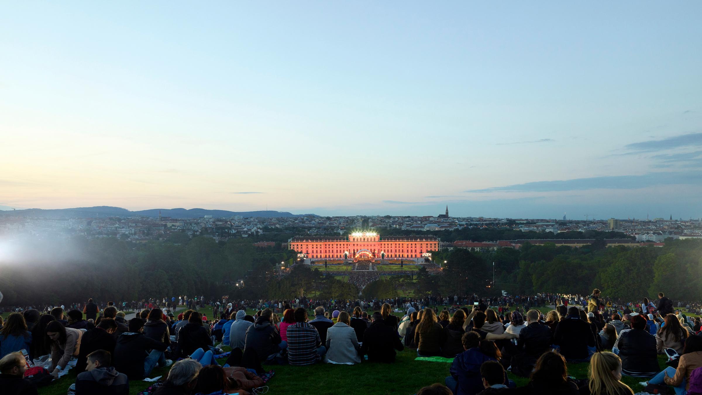 "Sommernachtskonzert Schönbrunn 2018": Frühabendstimmung:  Zu sehen ist der Schlosspark Schönbrunn mit tausenden von Leuten, die zu dem Sommernachtskonzert gekommen sind. Ganz hinten am Horizont ist das angestrahlte Schloss Schönbrunn mit einer Bühne zu erkennen.
