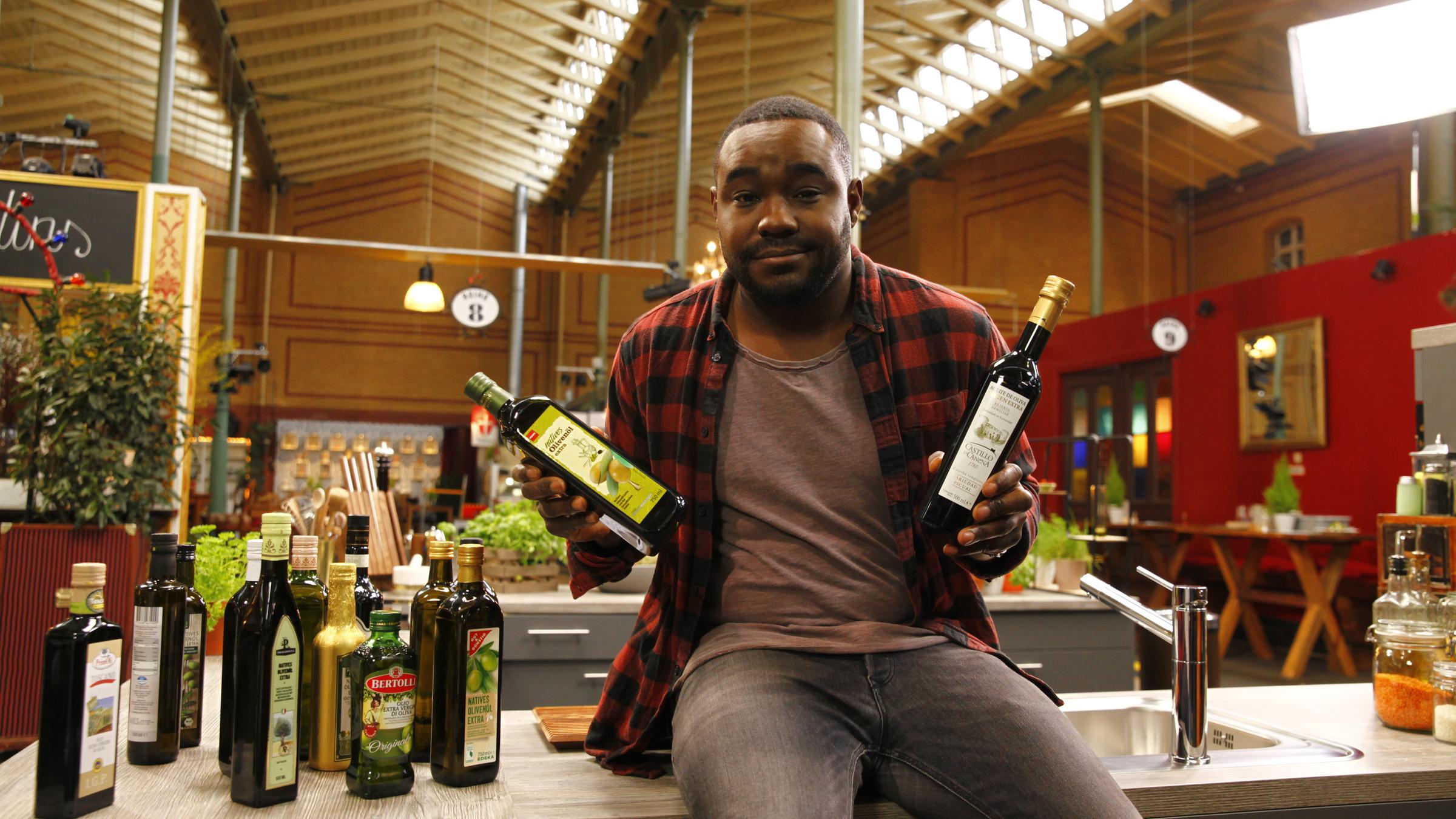 "ZDFzeit: Nelson Müllers Essens-Check - Wie gut sind Olivenöl, Lachs & Obst to go?": Nelson Müller hält zwei Flaschen in der Hand und schaut in die Kamera.