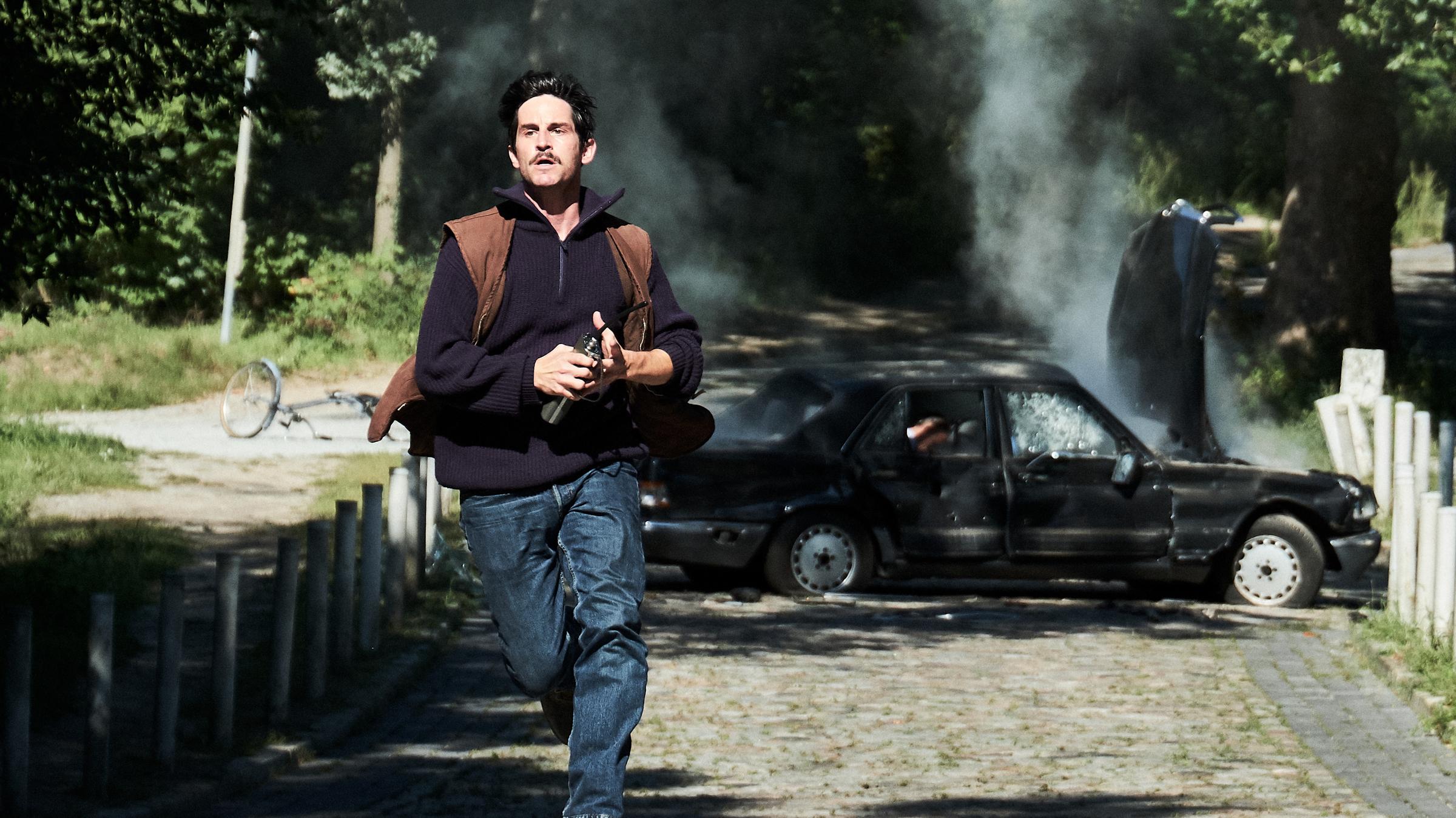 "Der Mordanschlag - Teil 1": Gelfert (Christoph Bach) rennt eine gepflasterte Straße entlang - schnellstens weg vom in die Luft gesprengtem Auto, das im Hintergrund rauchend quer über die Straße steht.