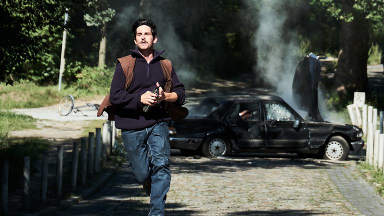 "Der Mordanschlag - Teil 1": Gelfert (Christoph Bach) rennt eine gepflasterte Straße entlang - schnellstens weg vom in die Luft gesprengtem Auto, das im Hintergrund rauchend quer über die Straße steht.