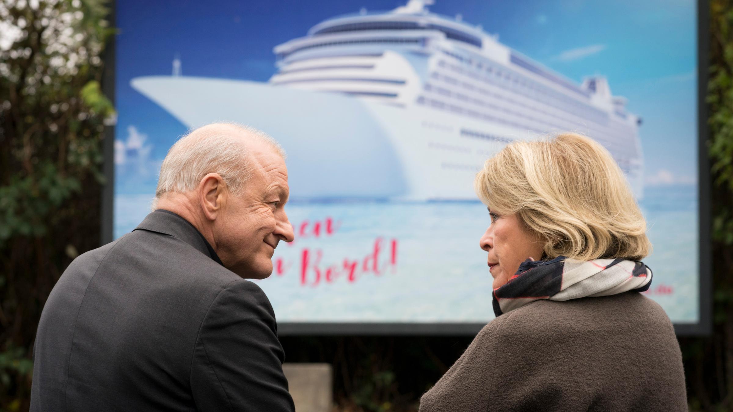 "Wilsberg - Mörderische Rendite": Georg Wilsberg (Leonard Lansink) und Anna Springer (Rita Russek) sitzen gemeinsam auf einer Bank vor einem Kreuzfahrt-Werbeplakat und schauen sich an.