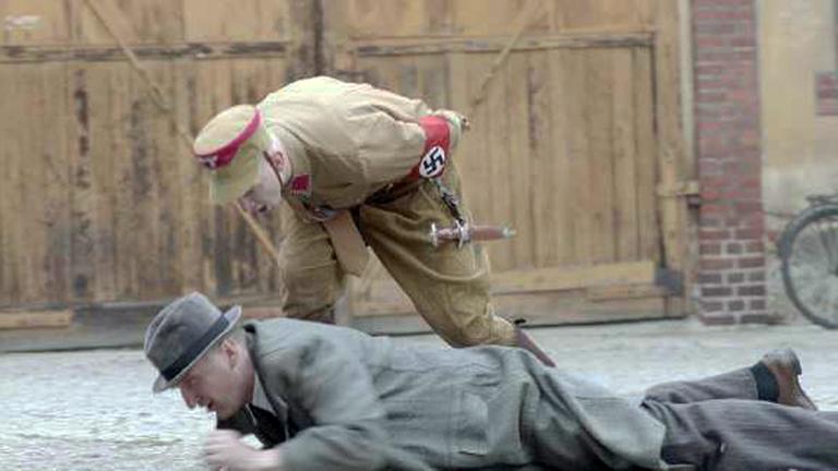 "ZDF-History: Verwüstet, zerstört, entrechtet: Das Novemberpogrom 1938": Auf Bauch liegender erwachsener Mann mit Hut wird von einem über ihm stehenden SA-Mann in Uniform angebrüllt. (Szenenfoto aus Dokumentation).