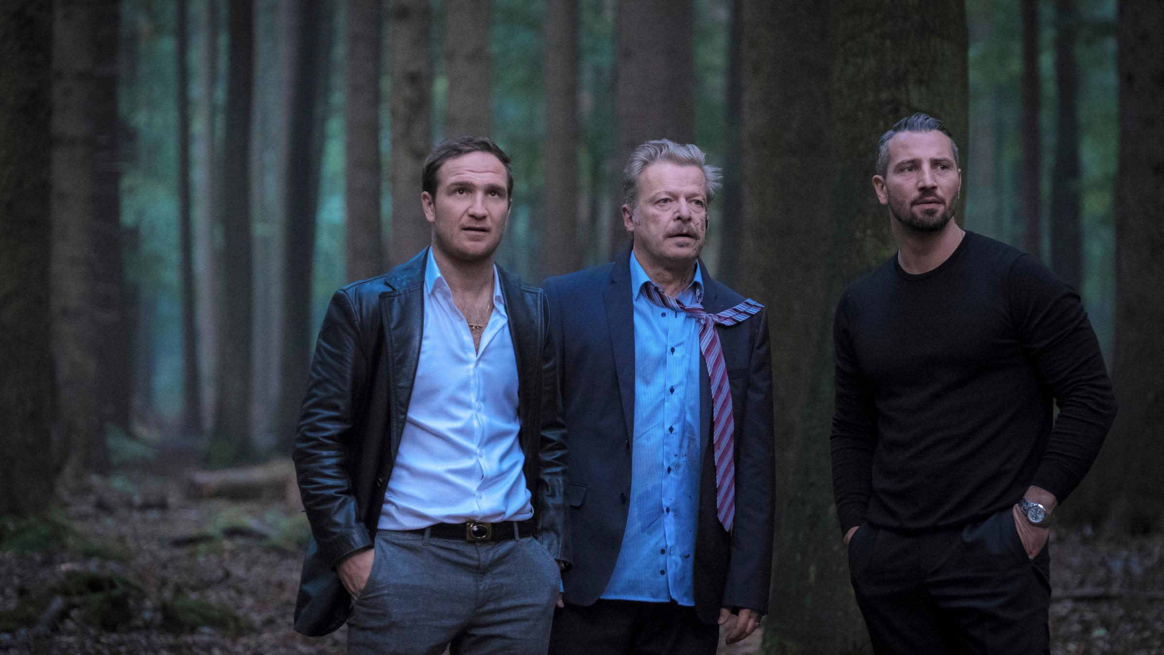 „Nachtschicht – Es lebe der Tod“: Angelo (Frederick Lau), Claude (Murathan Muslu)  und De Groot (Roland Koch) stehen nebeneinander im Wald und schauen in eine Richtung. De Groot sieht leicht verwahrlost aus, sein Hemd ist fleckig, und die Krawatte hängt über einer Schulter.