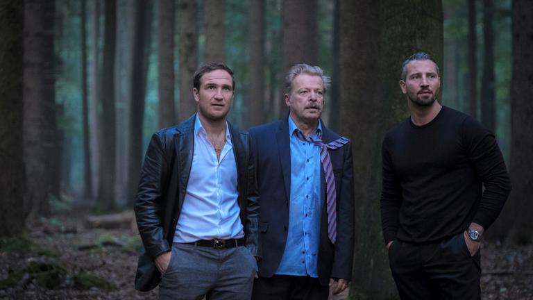 „Nachtschicht – Es lebe der Tod“: Angelo (Frederick Lau), Claude (Murathan Muslu)  und De Groot (Roland Koch) stehen nebeneinander im Wald und schauen in eine Richtung. De Groot sieht leicht verwahrlost aus, sein Hemd ist fleckig, und die Krawatte hängt über einer Schulter.