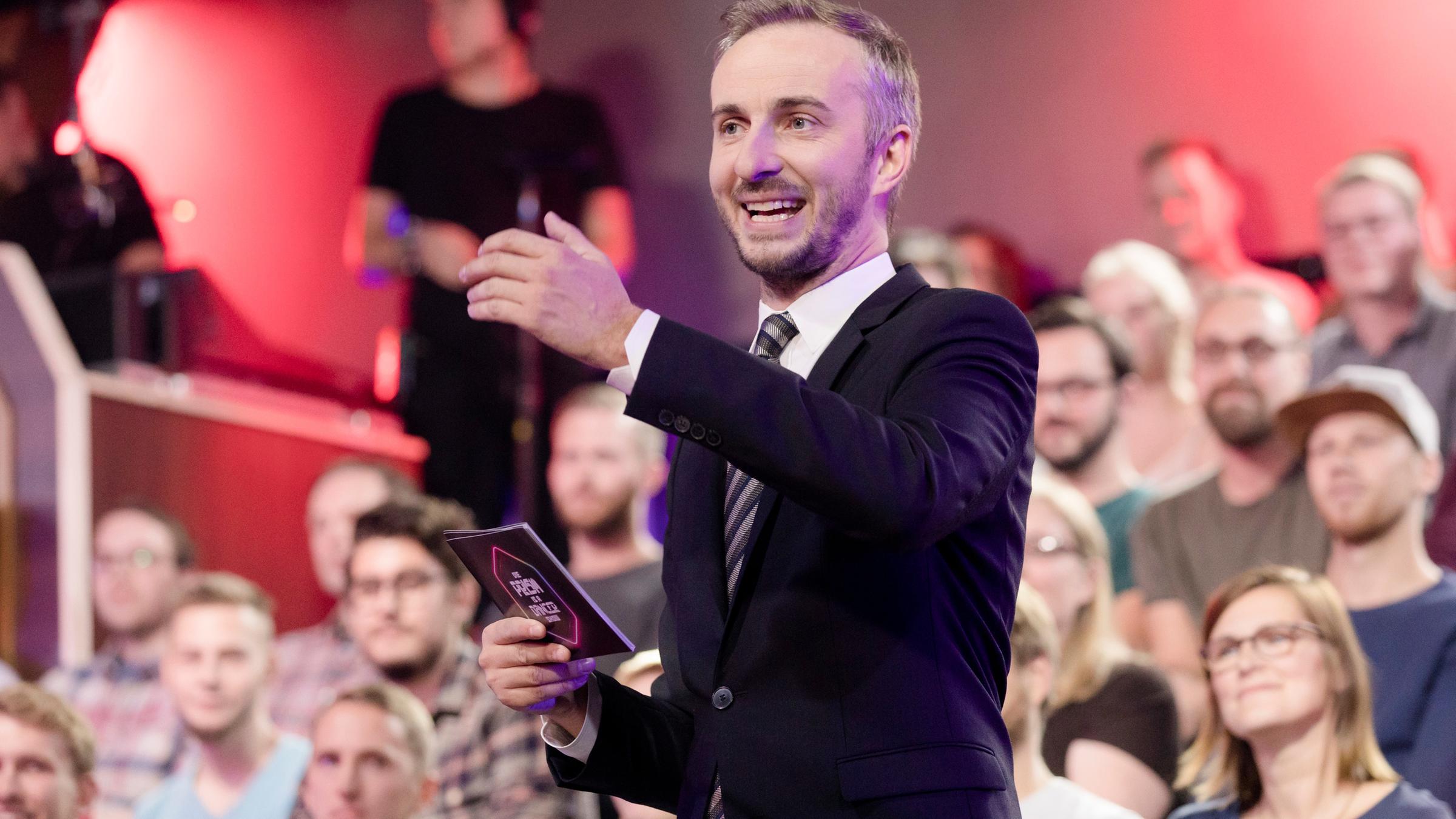 "Lass dich überwachen! Die PRISM IS A DANCER Show": Jan Böhmermann steht vor Studiopublikum. In der rechten Hand hält er eine Moderationskarte, die linke ist halbhoch erhoben.